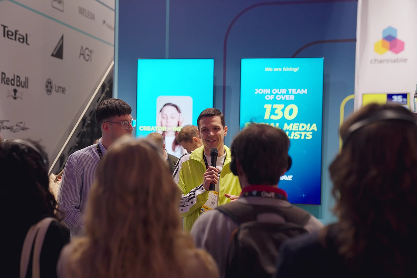 Ein Mann in leuchtend gelber Jacke spricht in ein Mikrofon vor Publikum bei einer Veranstaltung mit zwei Bildschirmen im Hintergrund, die eine Social-Media-Stellenanzeige zeigen.