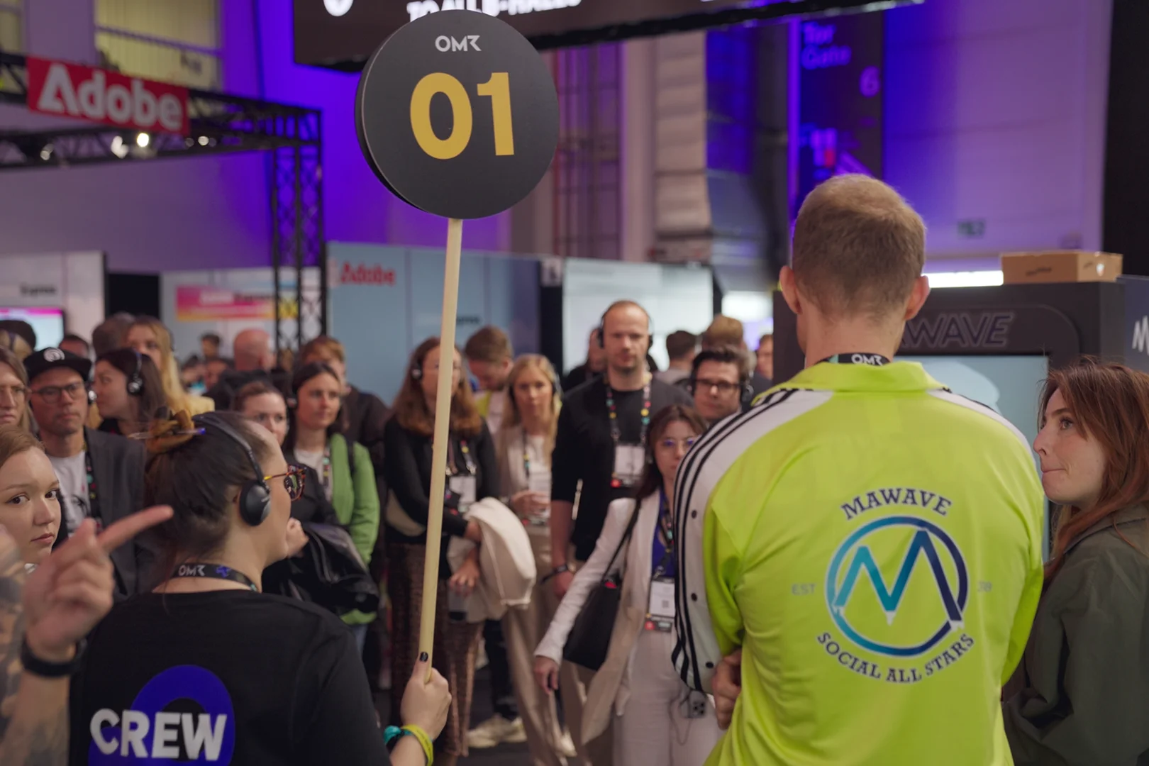 Menschenmenge bei einer Veranstaltung, eine Person hält ein Schild mit der Nummer 01, im Hintergrund ein Adobe-Logo.