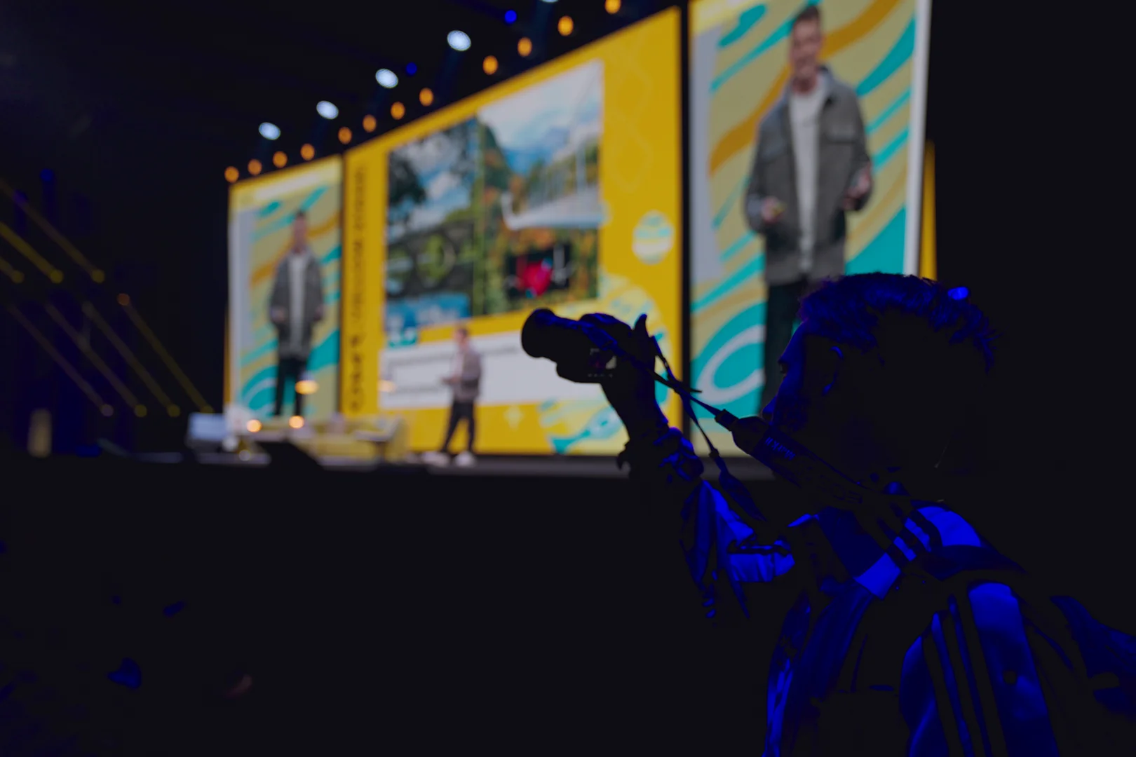 Silhouette einer Person mit Kamera im Vordergrund bei einer Präsentation auf einer Bühne mit großem Bildschirm im Hintergrund.