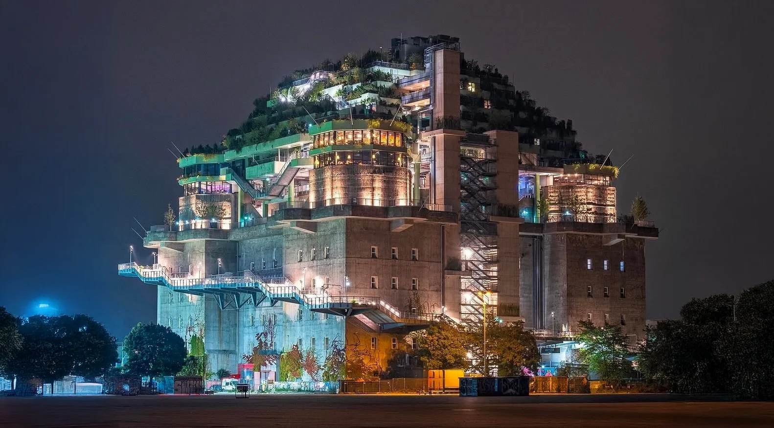 Beleuchteter mehrstöckiger Bunker mit begrünten Terrassen und moderner Architektur bei Nacht.