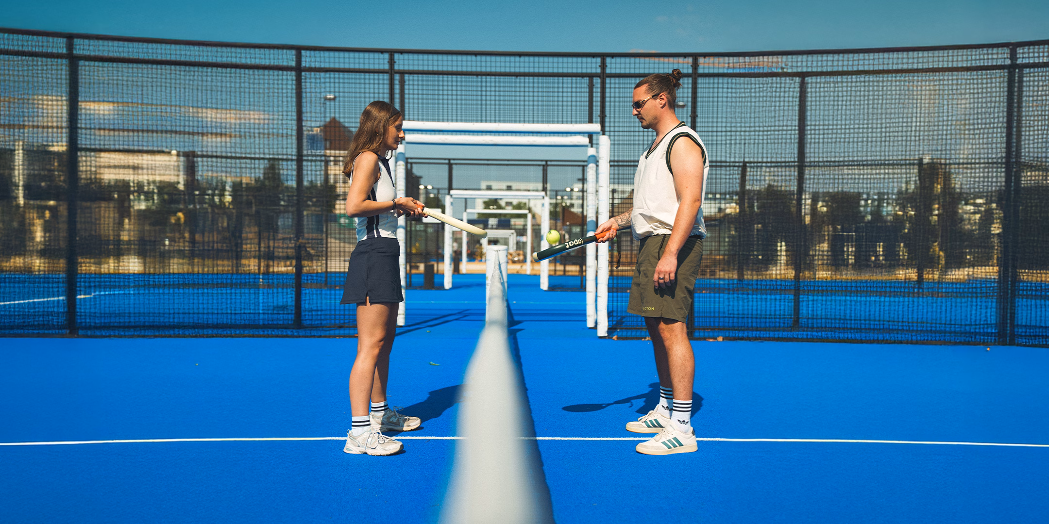 Zwei junge Erwachsene spielen Paddle-Tennis auf blauem Platz vor Netz an sonnigem Tag.