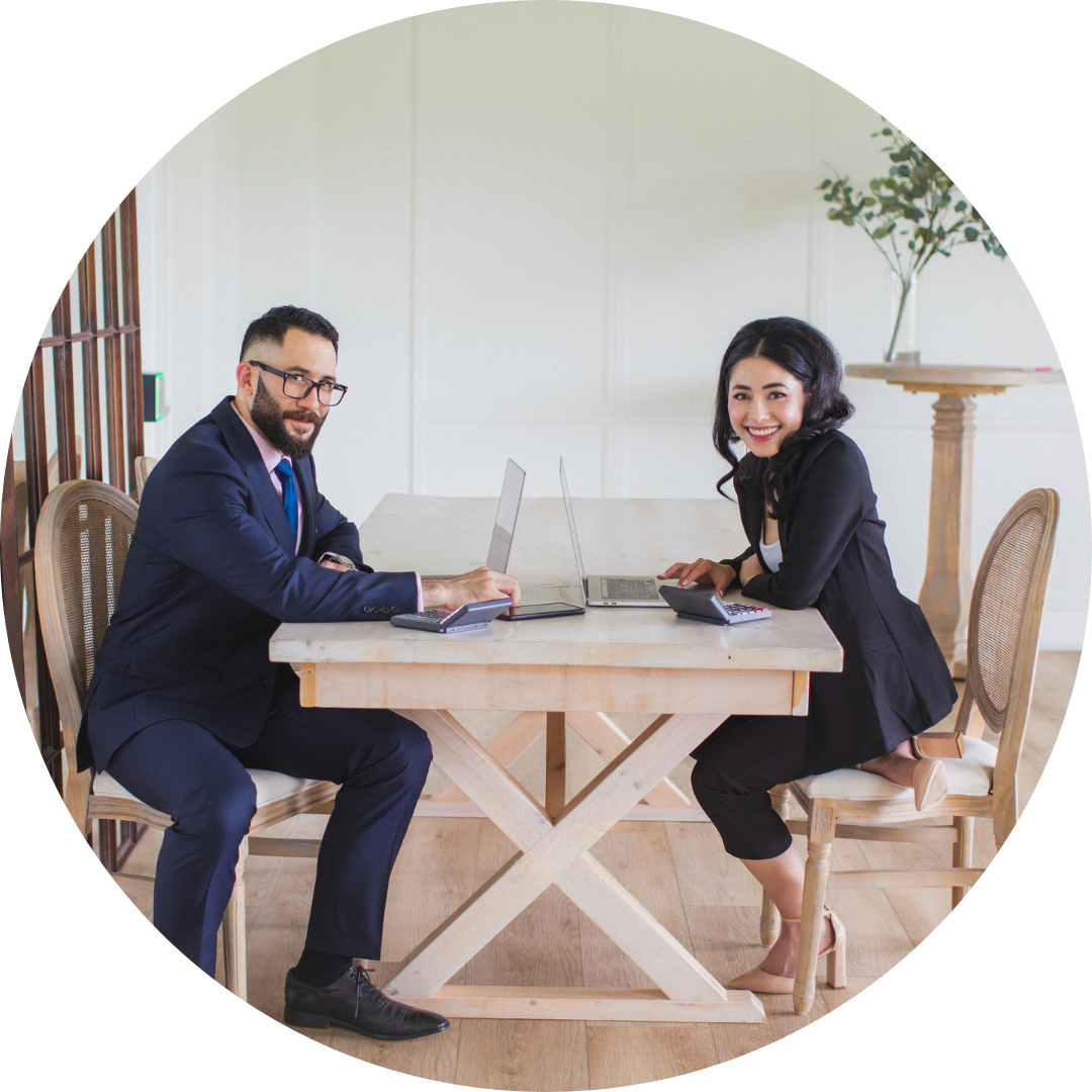 A man and a woman in business attire sitting at a wooden table with laptops and calculators, smiling at the camera.