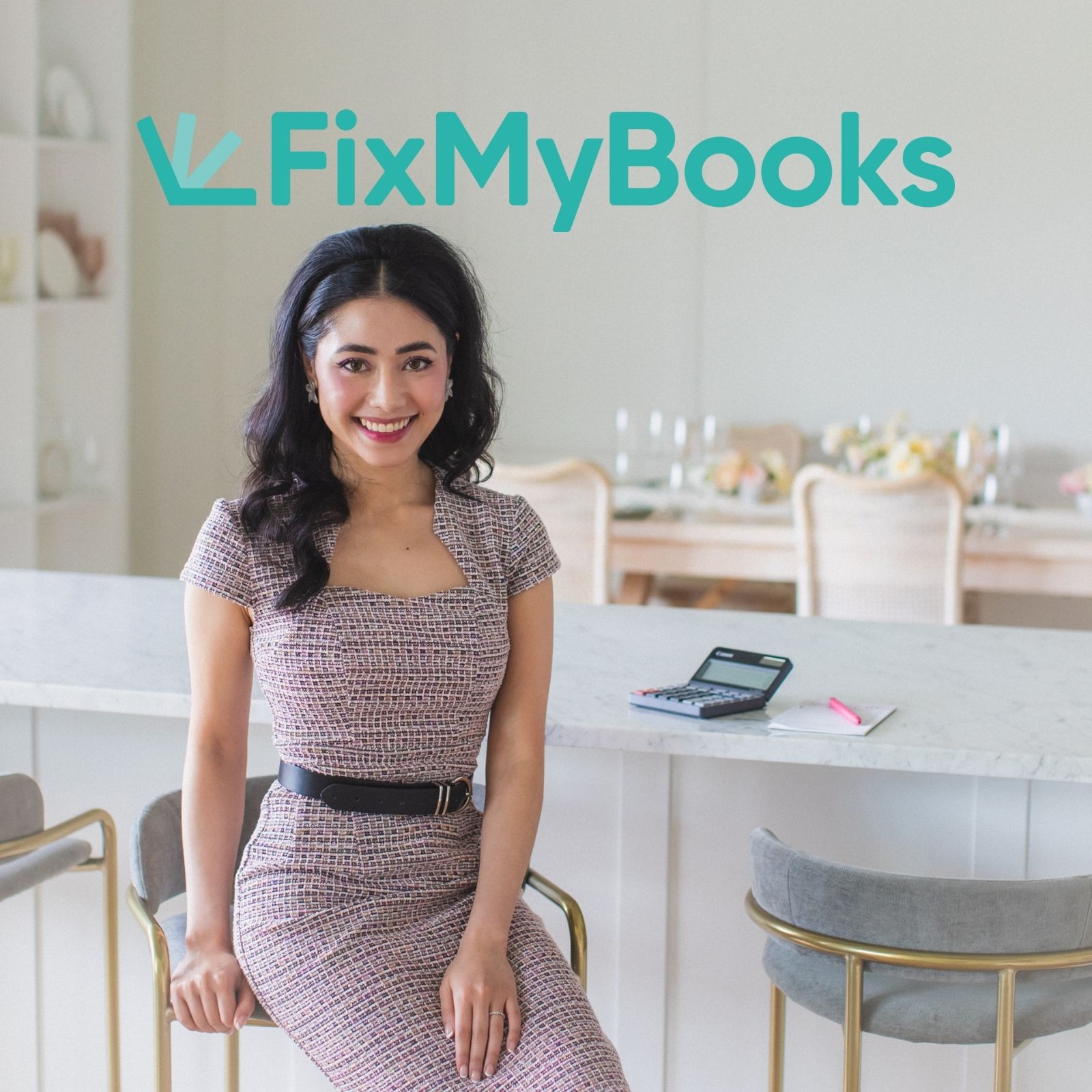 Smiling woman in a patterned dress sitting on a modern chair at a kitchen island with a calculator and notebook.