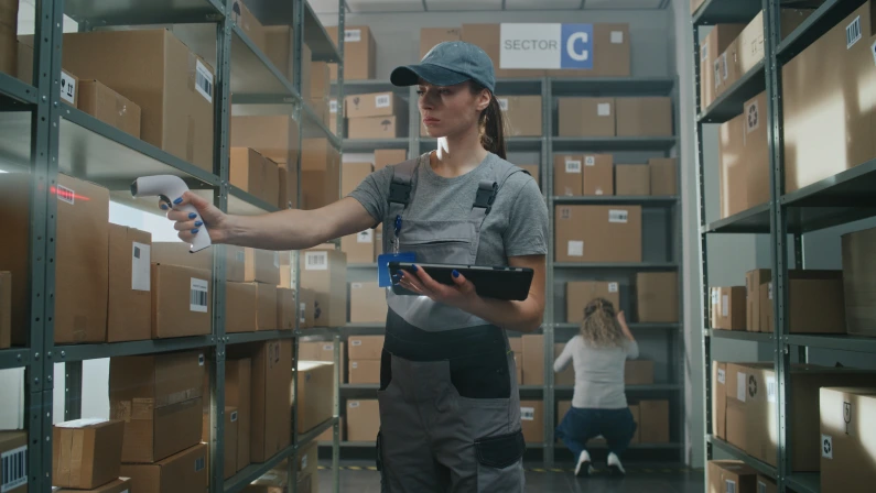 High-Tech Advanced Warehouse: Female Manager Scans Parcels, Checks Inventory on Tablet Computer. Worker in Futuristic Full Body Powered Exoskeleton with Heavy Cardboard Box Walks in the Background.