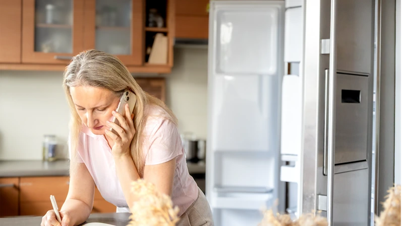 Woman talks to repair technician on phone and writes appointment details in notebook. Appliance service scheduling, household problem solving and maintenance planning concept