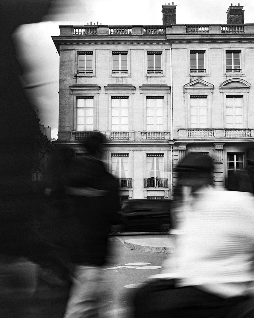 Black and white photo of a stately building with blurred figures moving in the foreground, contrasting stillness and motion.