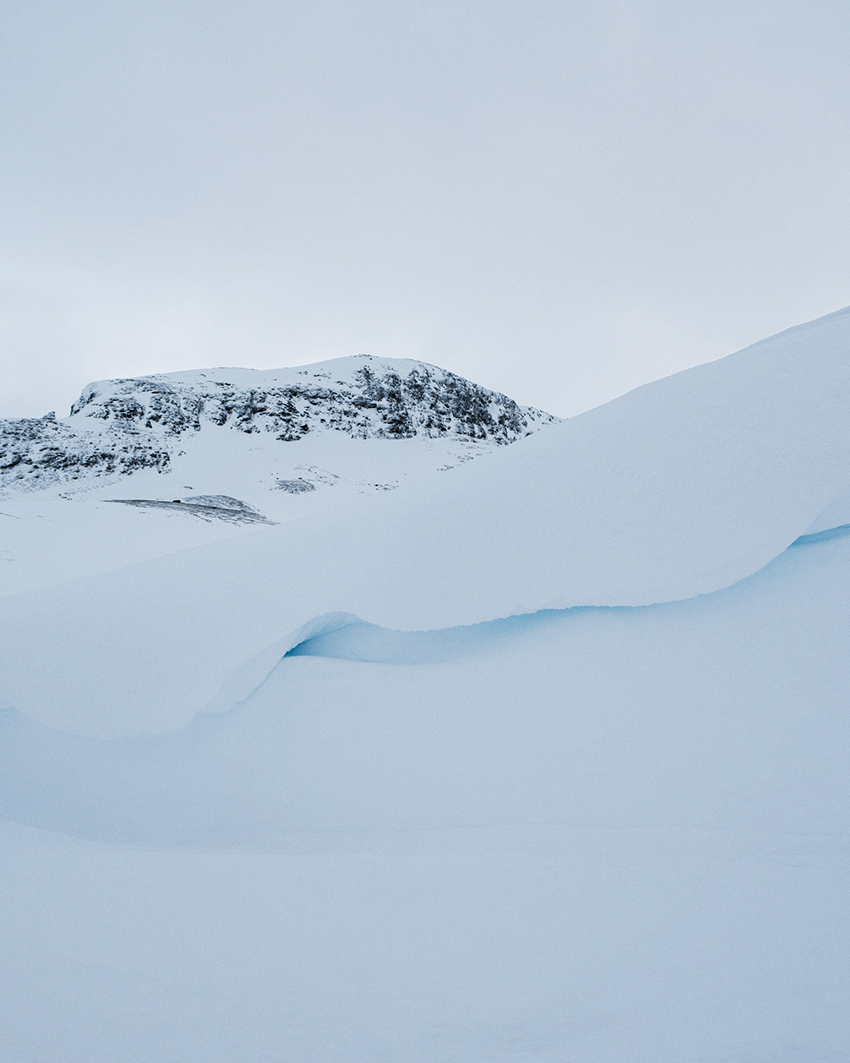 Snow-covered mountain peak in Norway with blue-tinted snow formations in the foreground, captured in a minimalist style.