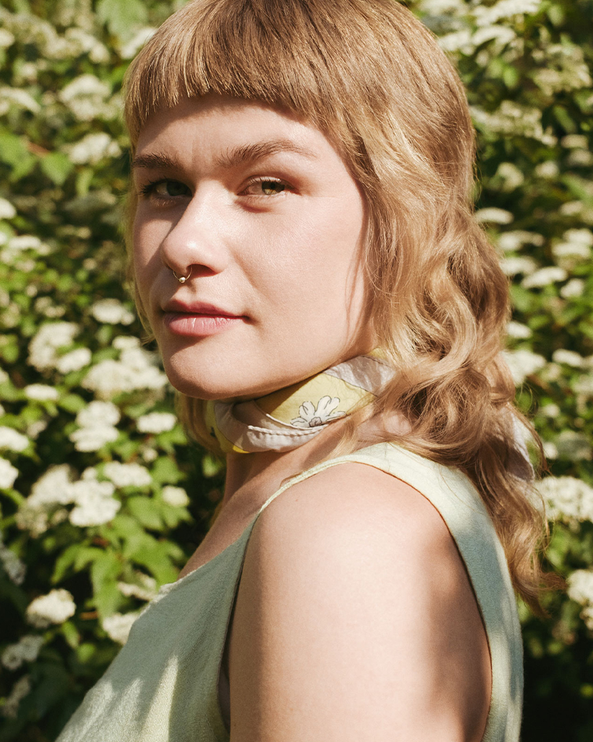 Close-up portrait of Katinka Askehave with light hair and scarf around her neck, standing among blooming white flowers and looking toward the camera.
