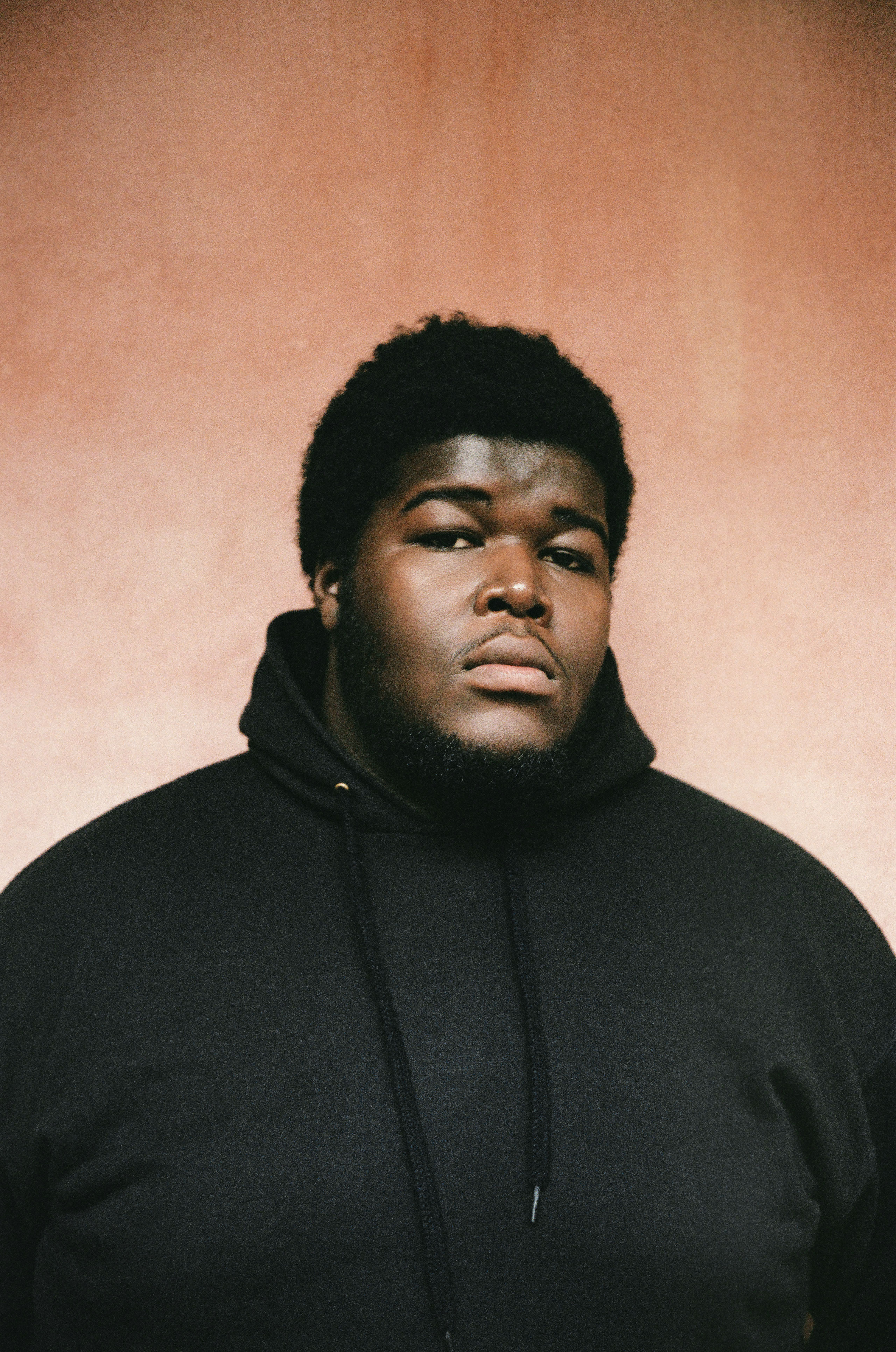 Studio portrait of Danish artist BUKURU wearing a black hoodie against a warm textured background.