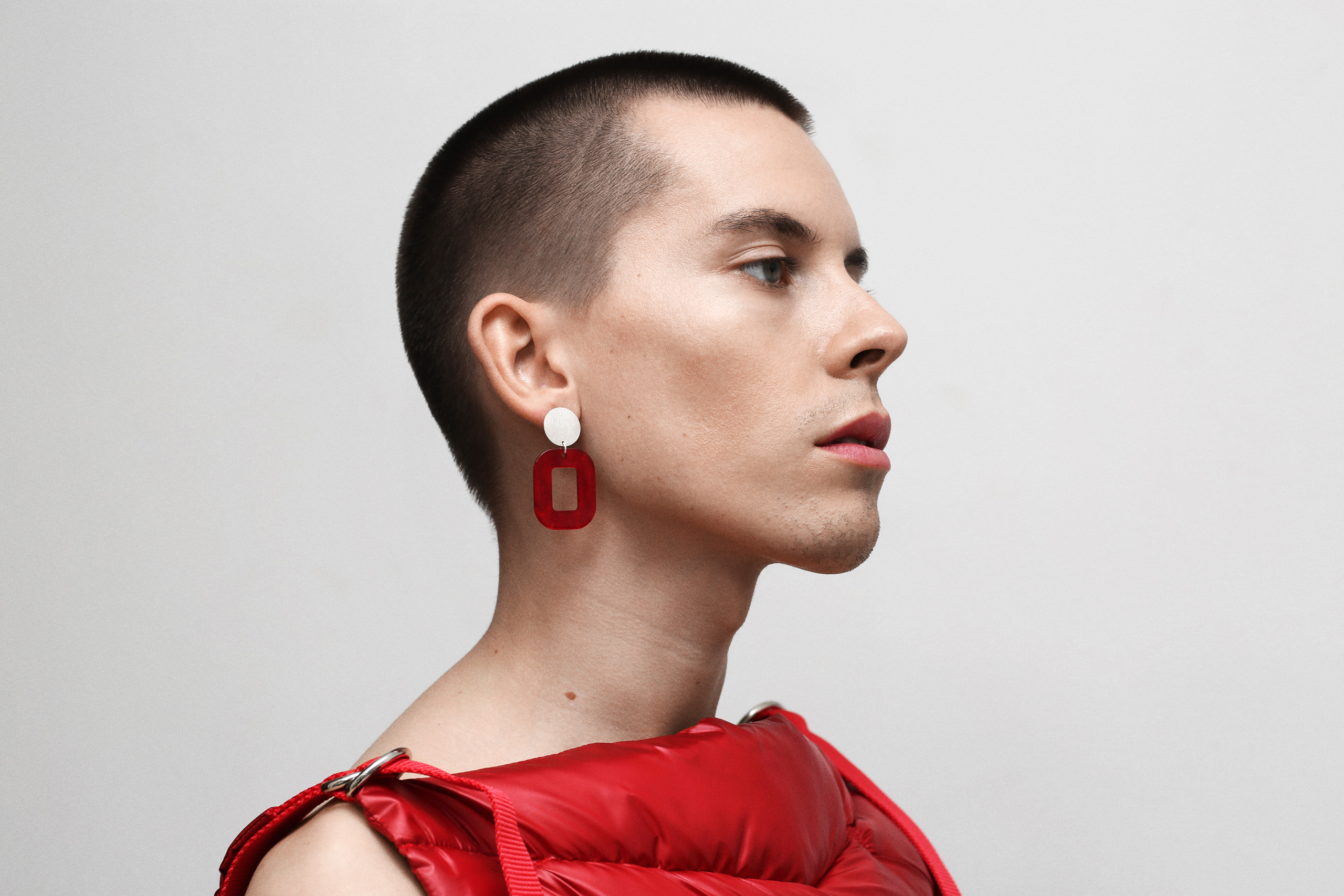 Portrait of Emil Ljungberg wearing a red Persuede Copenhagen garment and statement earrings photographed in a minimalist studio setting.