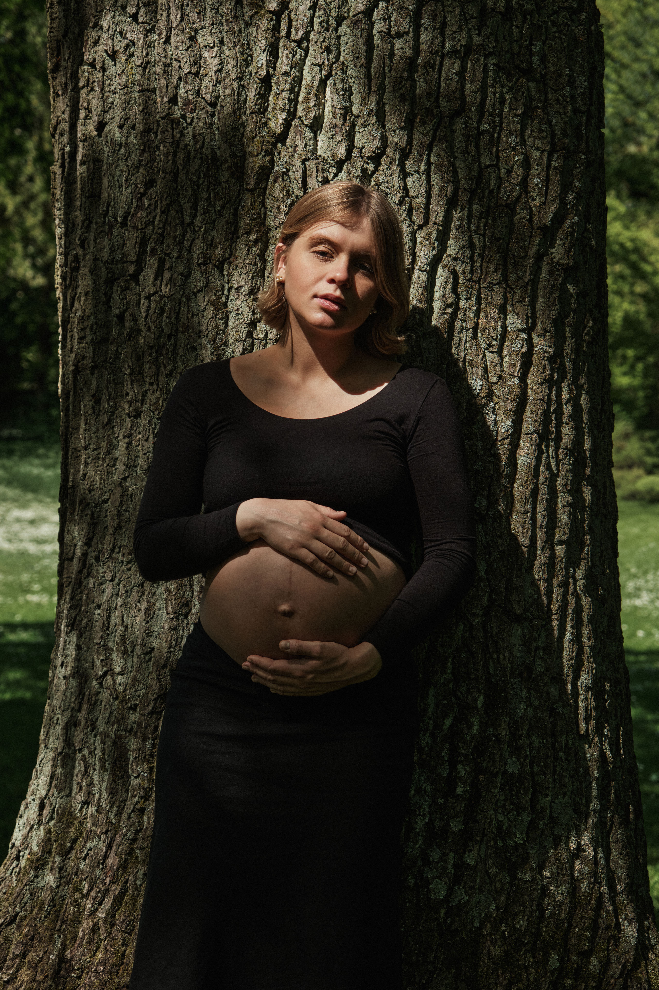     Maternity portrait of Jana Katrín standing against a large tree in a sunlit garden in Copenhagen.
