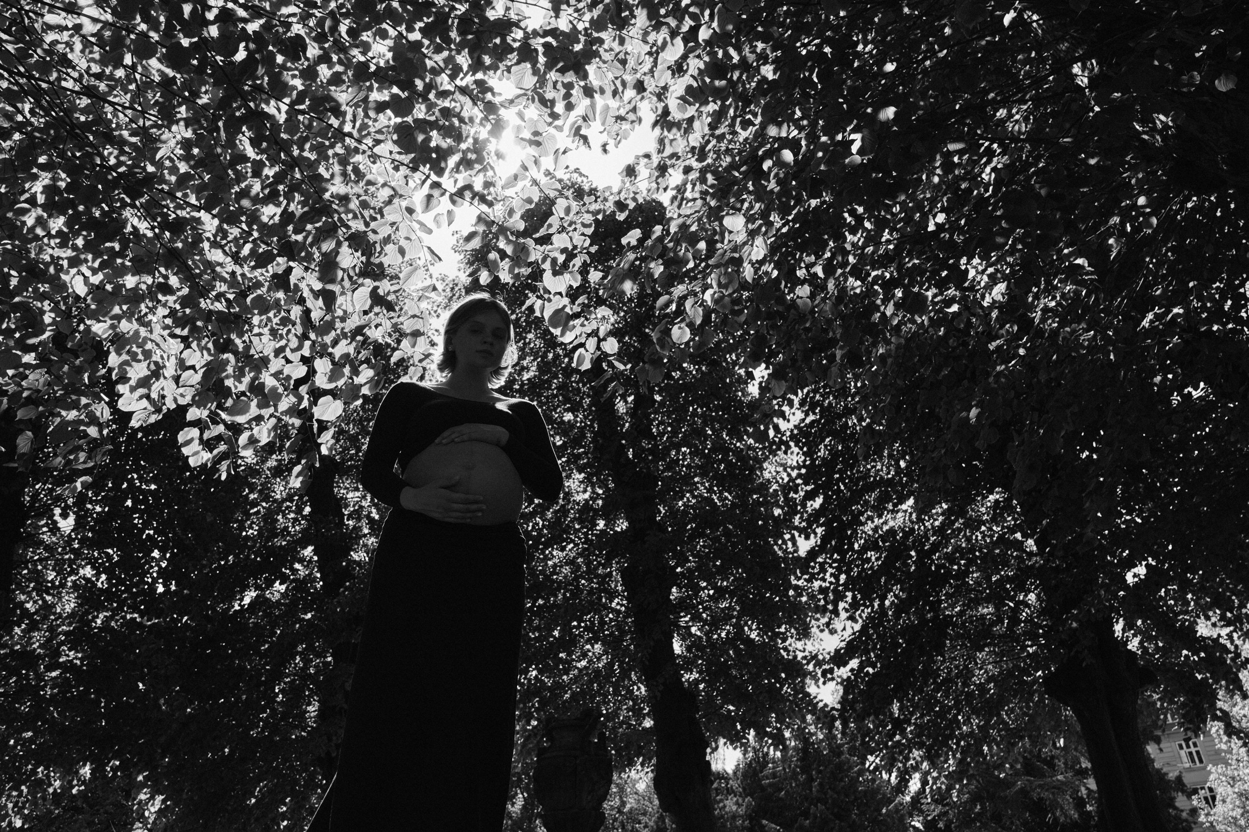     Black and white maternity portrait of Jana Katrín standing beneath a dense canopy of sunlit leaves in Copenhagen.