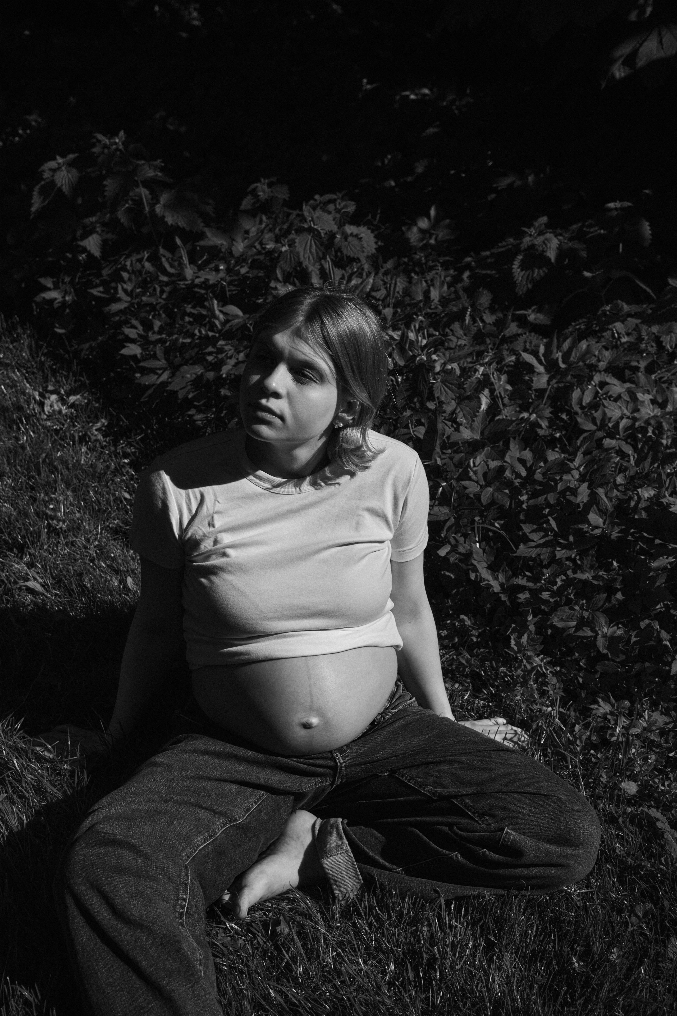    Black and white maternity portrait of Jana Katrín sitting on the grass in dramatic sunlight in a Copenhagen garden.