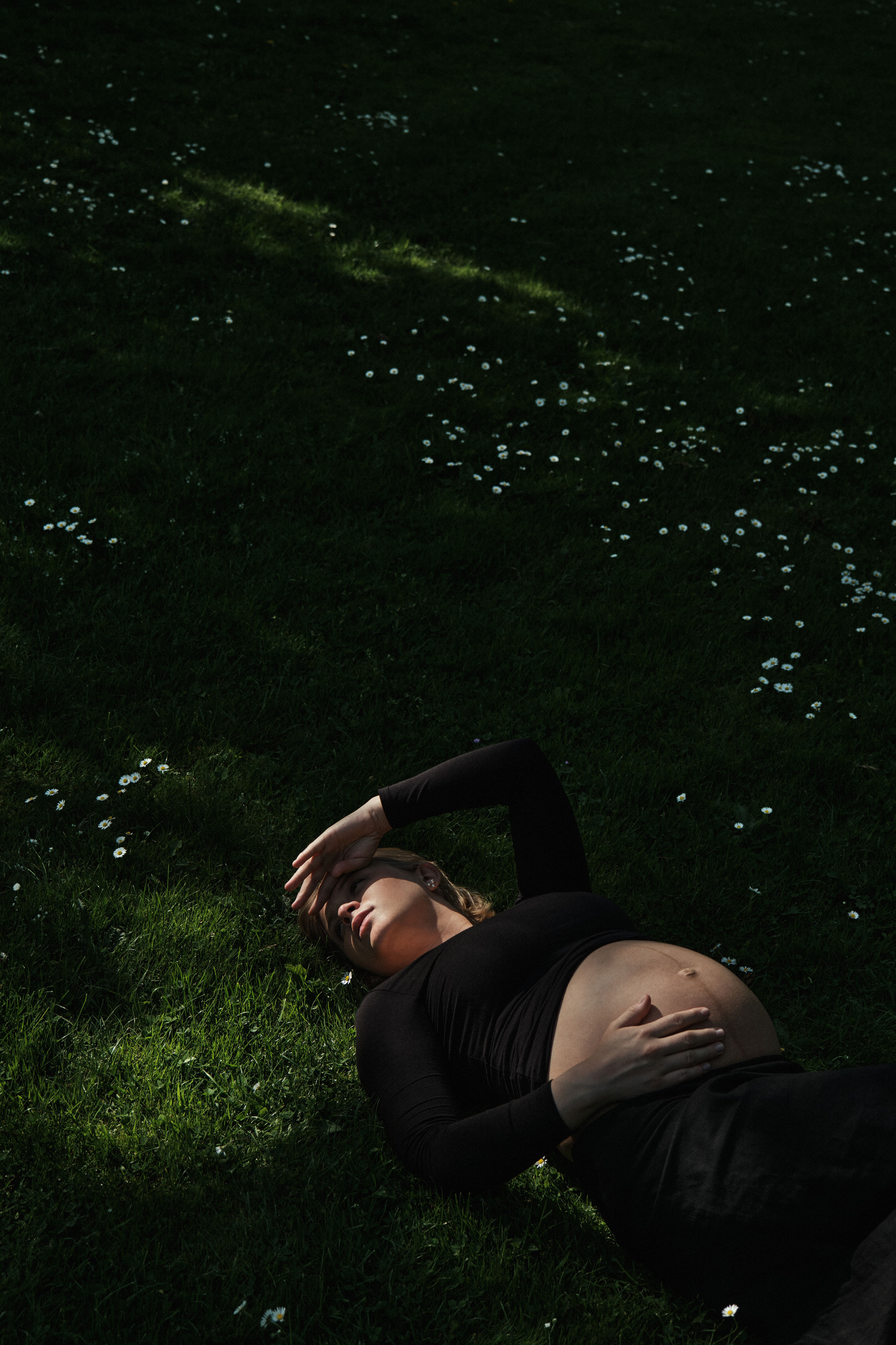     Jana Katrín lying in the grass among small white flowers in an outdoor maternity portrait in Copenhagen.