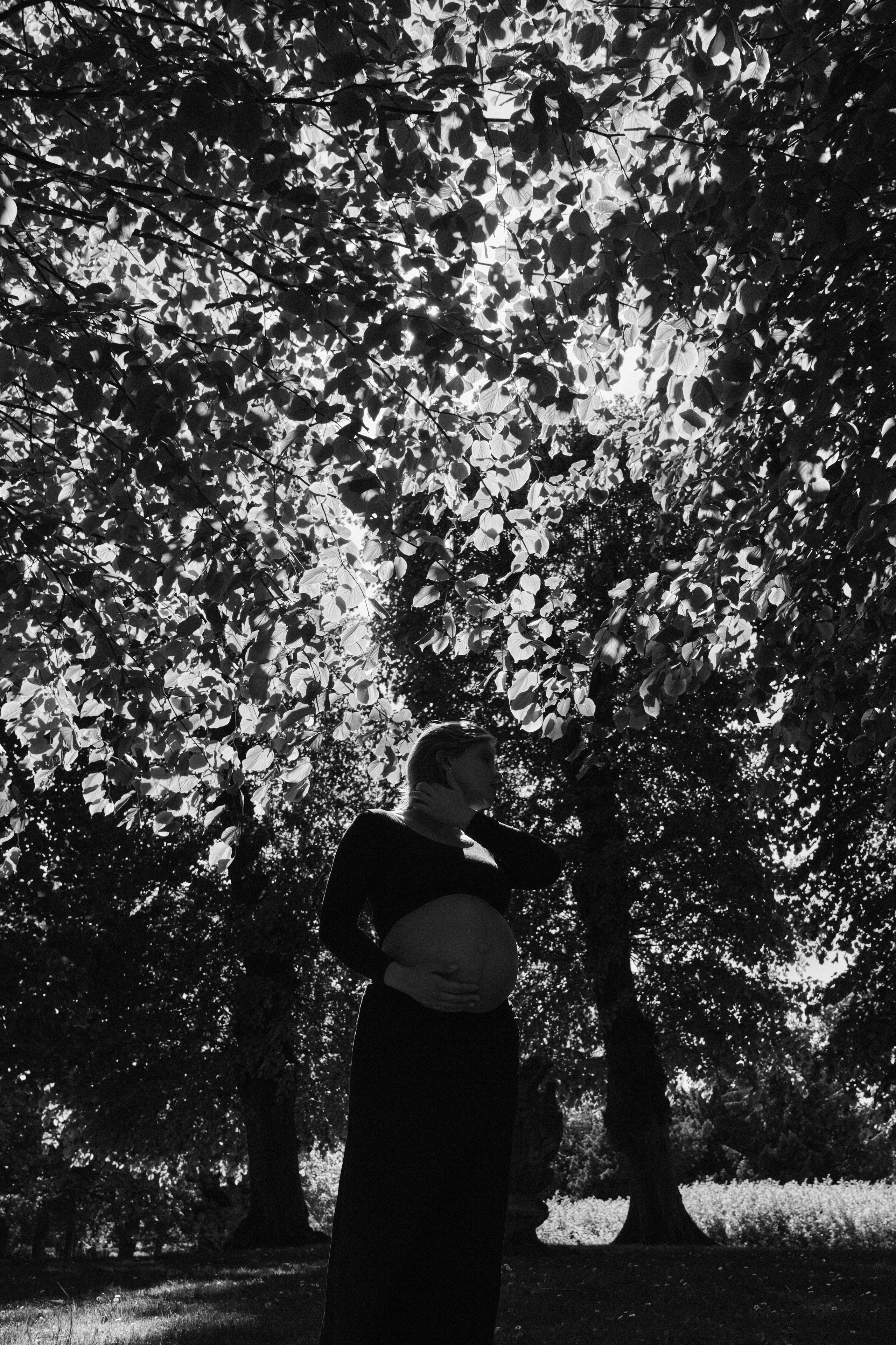     Black and white maternity photograph of Jana Katrín standing beneath trees and holding her pregnant belly in a Copenhagen garden.