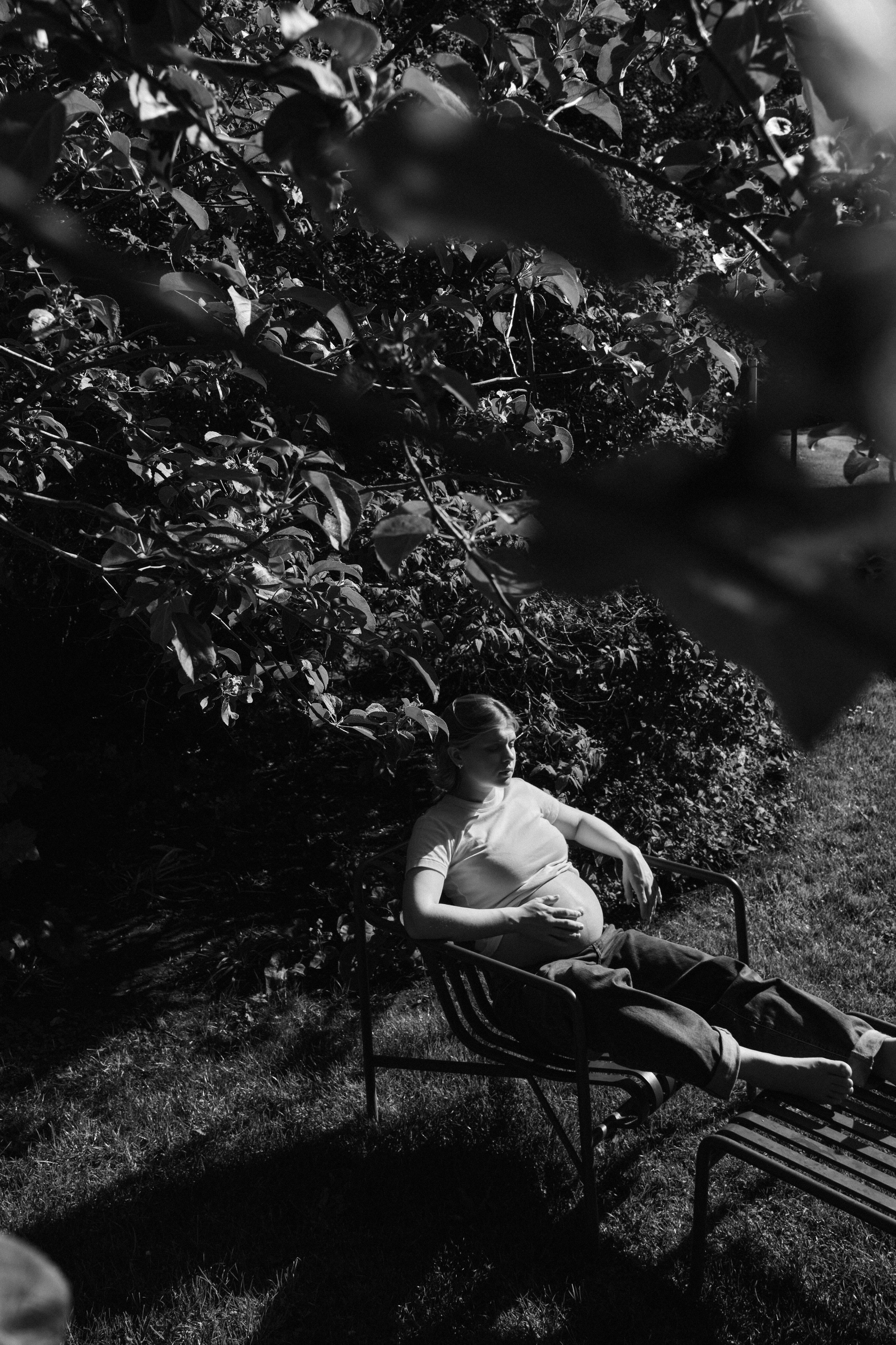     Black and white maternity photograph of Jana Katrín reclining on a garden chair beneath leafy branches in Copenhagen.