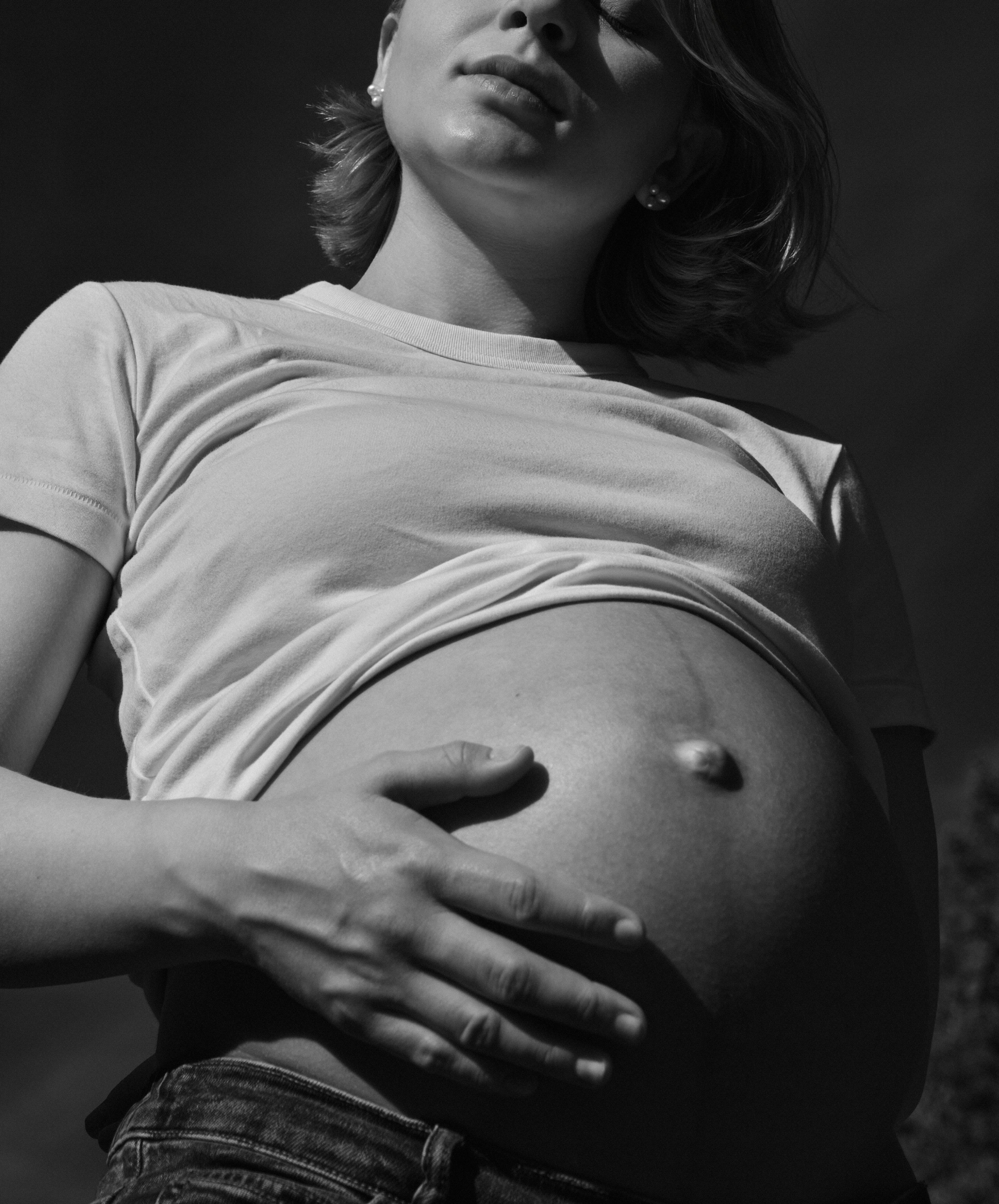     Close-up black and white maternity photograph of Jana Katrín holding her pregnant belly in direct sunlight.