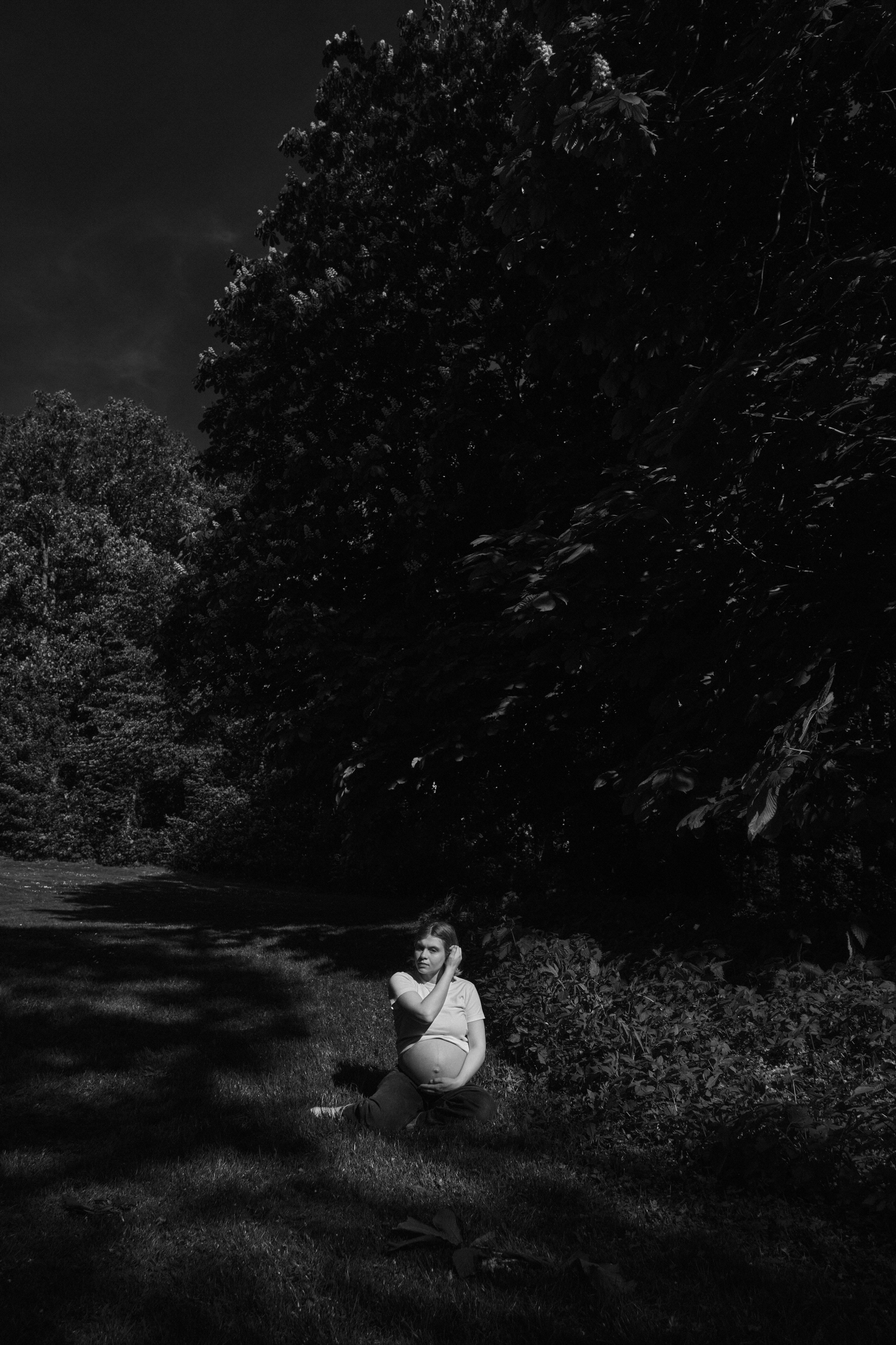     Black and white maternity photograph of Jana Katrín sitting in a shadowed garden landscape in Copenhagen.