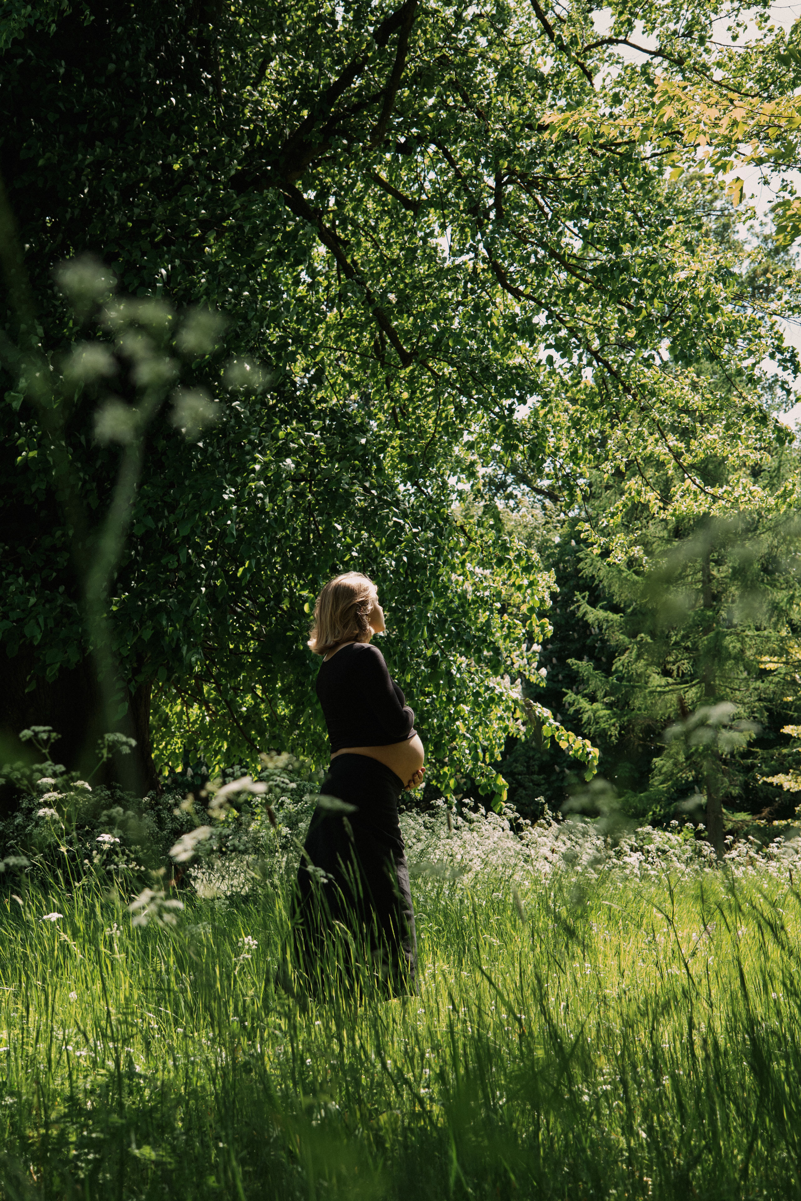     Jana Katrín standing in a sunlit meadow surrounded by lush greenery during an outdoor maternity photoshoot.