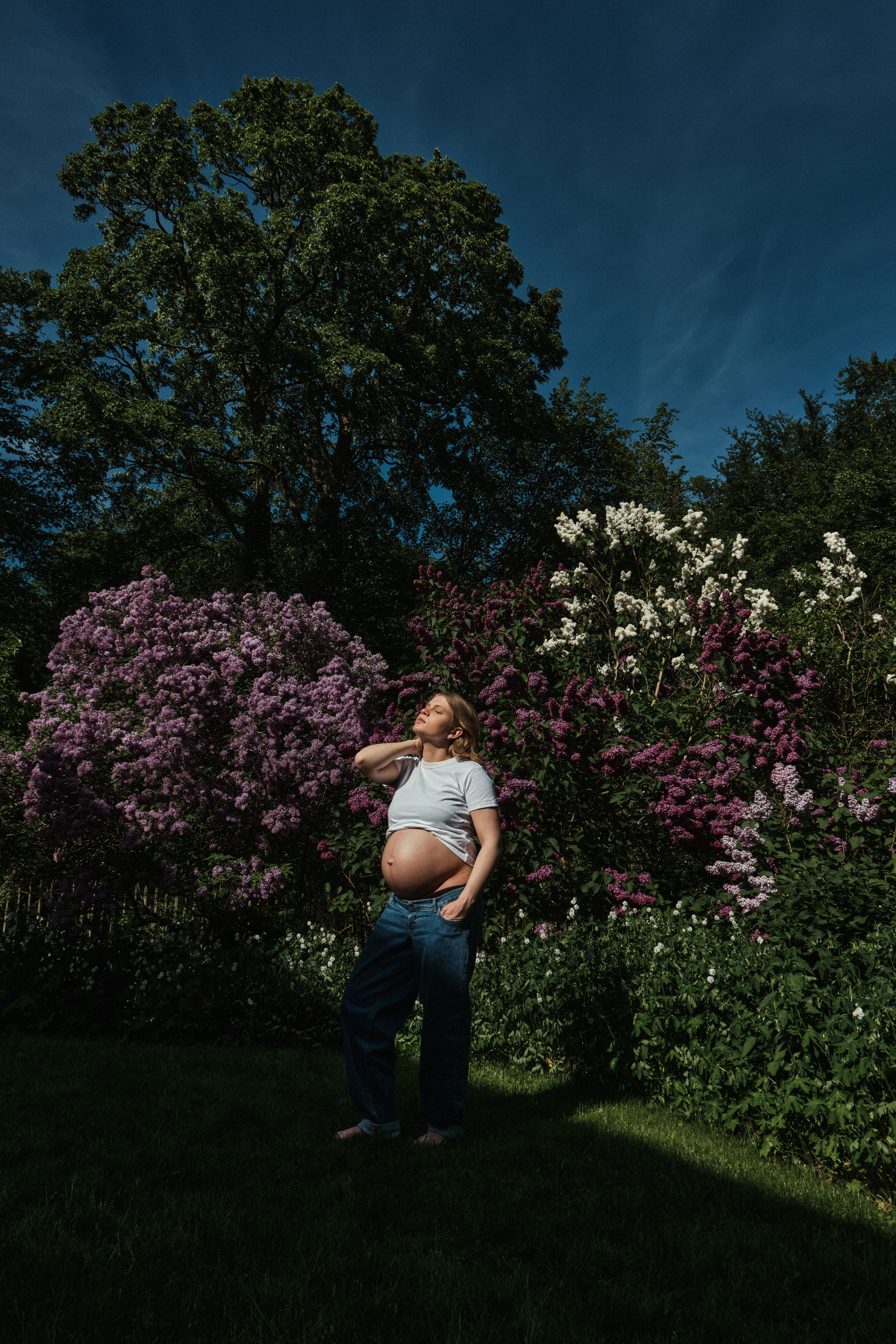     Maternity portrait of Jana Katrín standing in front of flowering lilac bushes beneath a deep blue sky.