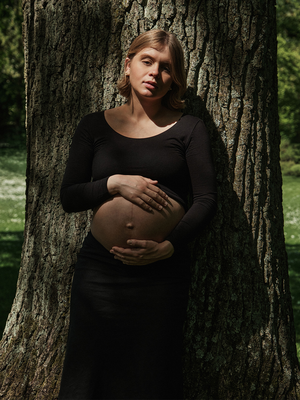 Maternity portrait of Jana Katrín standing between two large tree trunks in a Copenhagen garden.