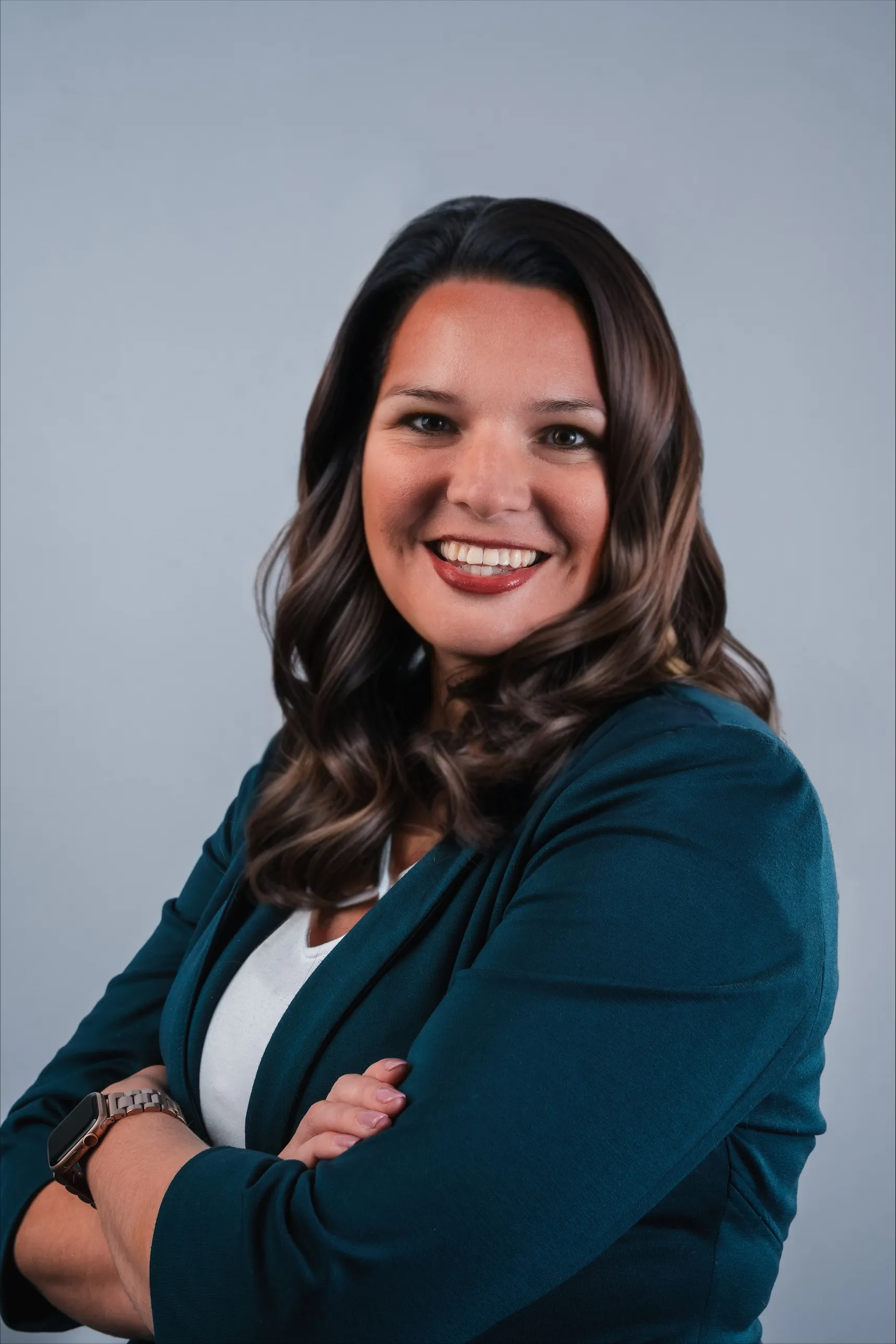 Smiling woman with shoulder-length wavy brown hair wearing a teal blazer and white top, arms crossed.