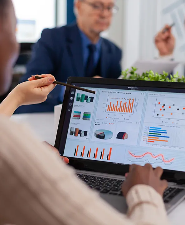 Person using a laptop showing various colorful graphs and charts, while another person points at the screen with a pen in a business meeting.