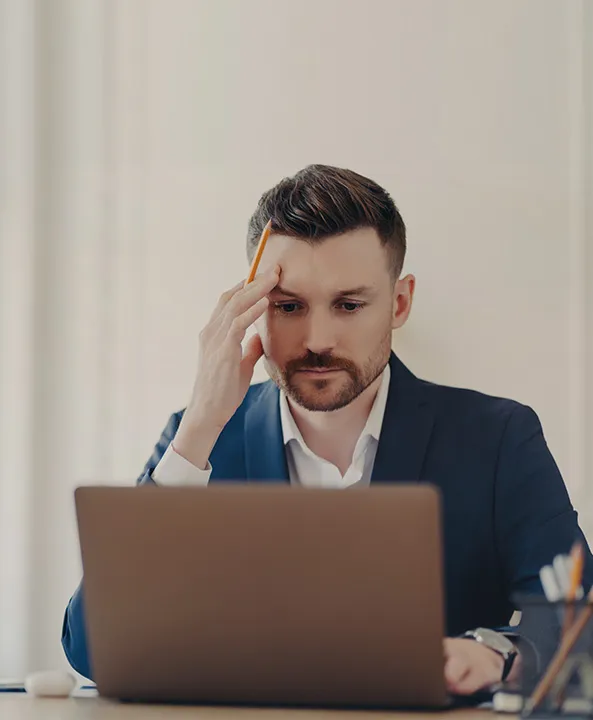 Man in a blue suit holding a pencil to his temple, concentrating on a laptop screen.