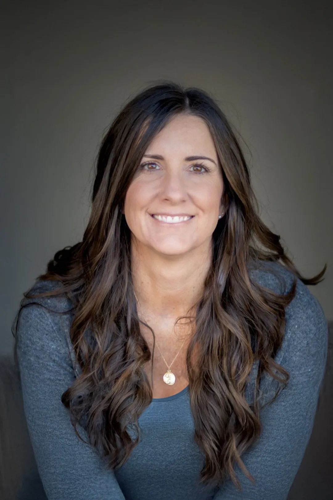 Smiling woman with long brown wavy hair wearing a blue top and gold pendant necklace against a plain background.