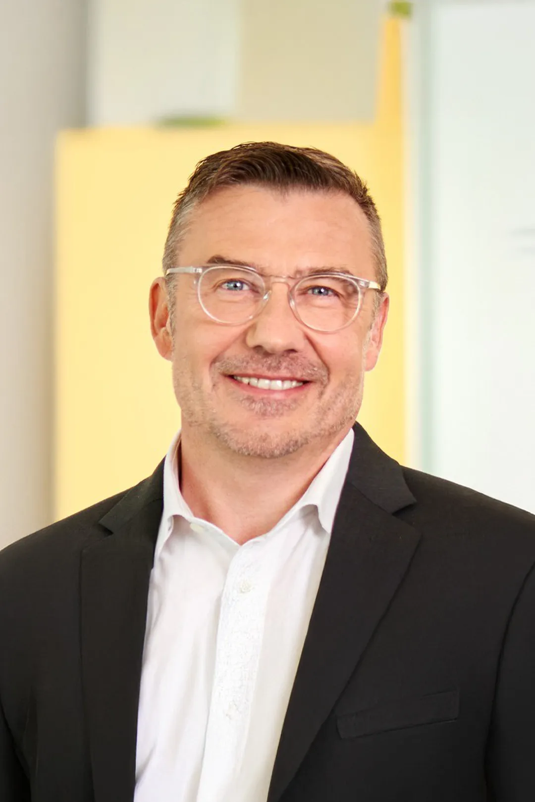 Smiling middle-aged man with short hair and glasses wearing a black suit jacket and white shirt in an indoor setting.