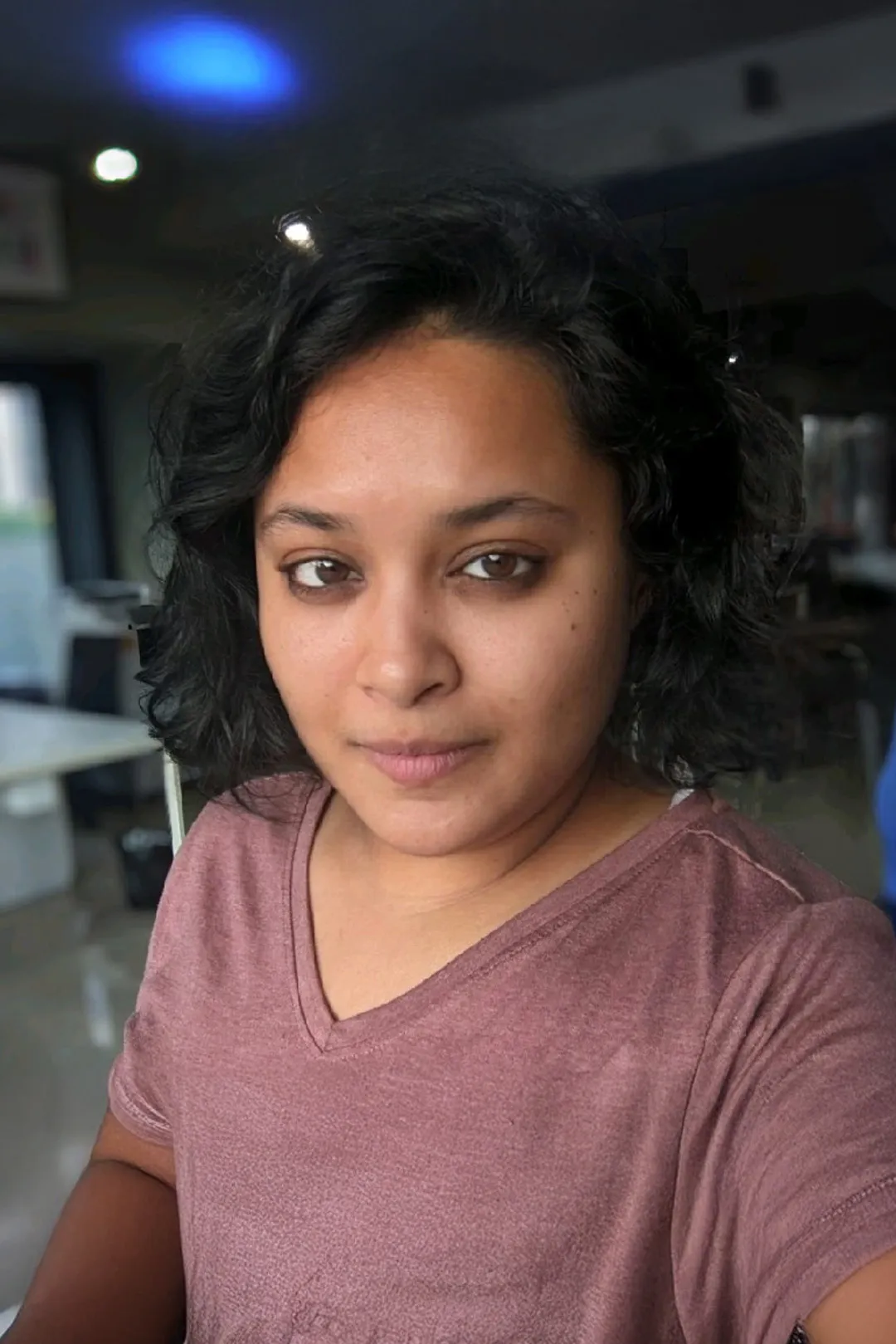 Woman with short curly hair wearing a mauve V-neck shirt looking at the camera indoors.