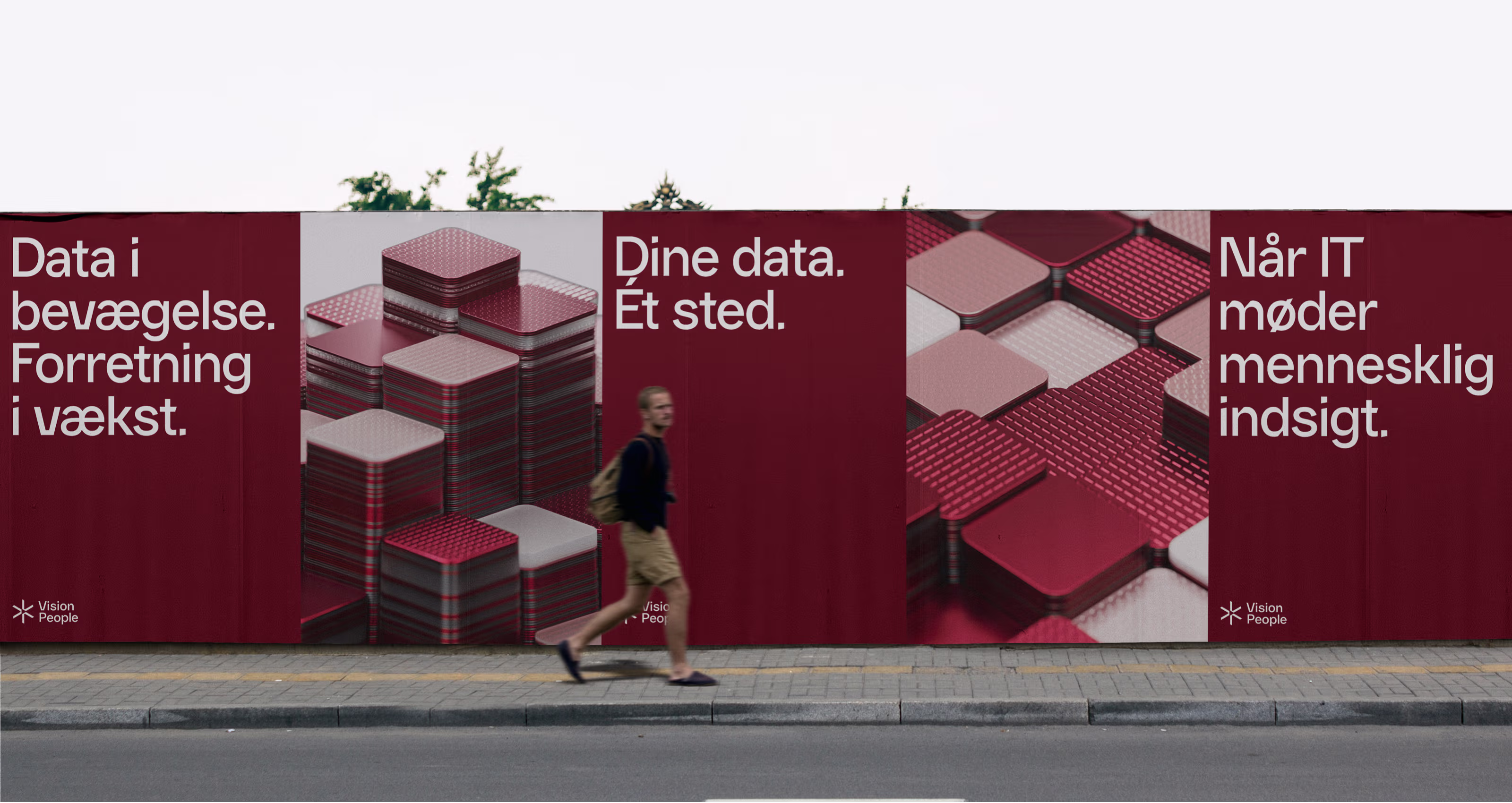 A person walking on a sidewalk in front of a large red urban poster with abstract stacked squares and texts about data and IT in Danish.