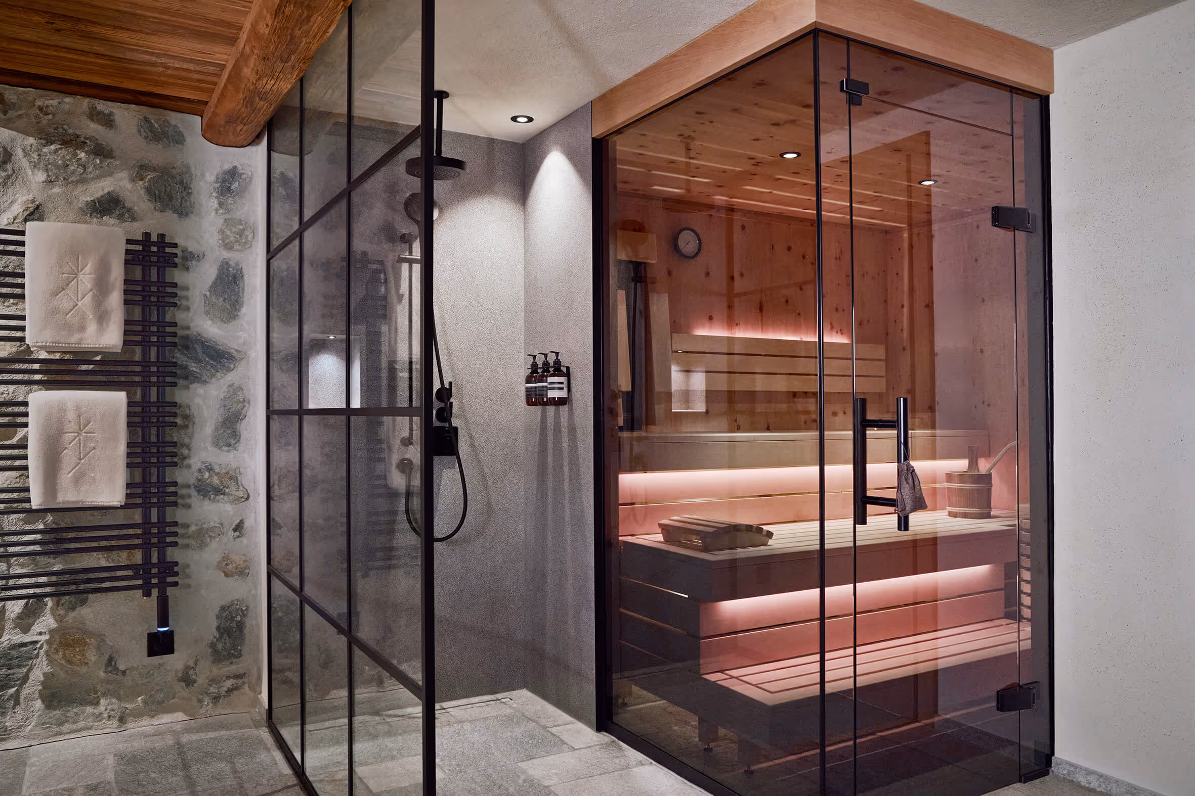 Moderne Sauna mit Glastür und angrenzender Dusche in einem Badezimmer mit Steinwand und Handtüchern.