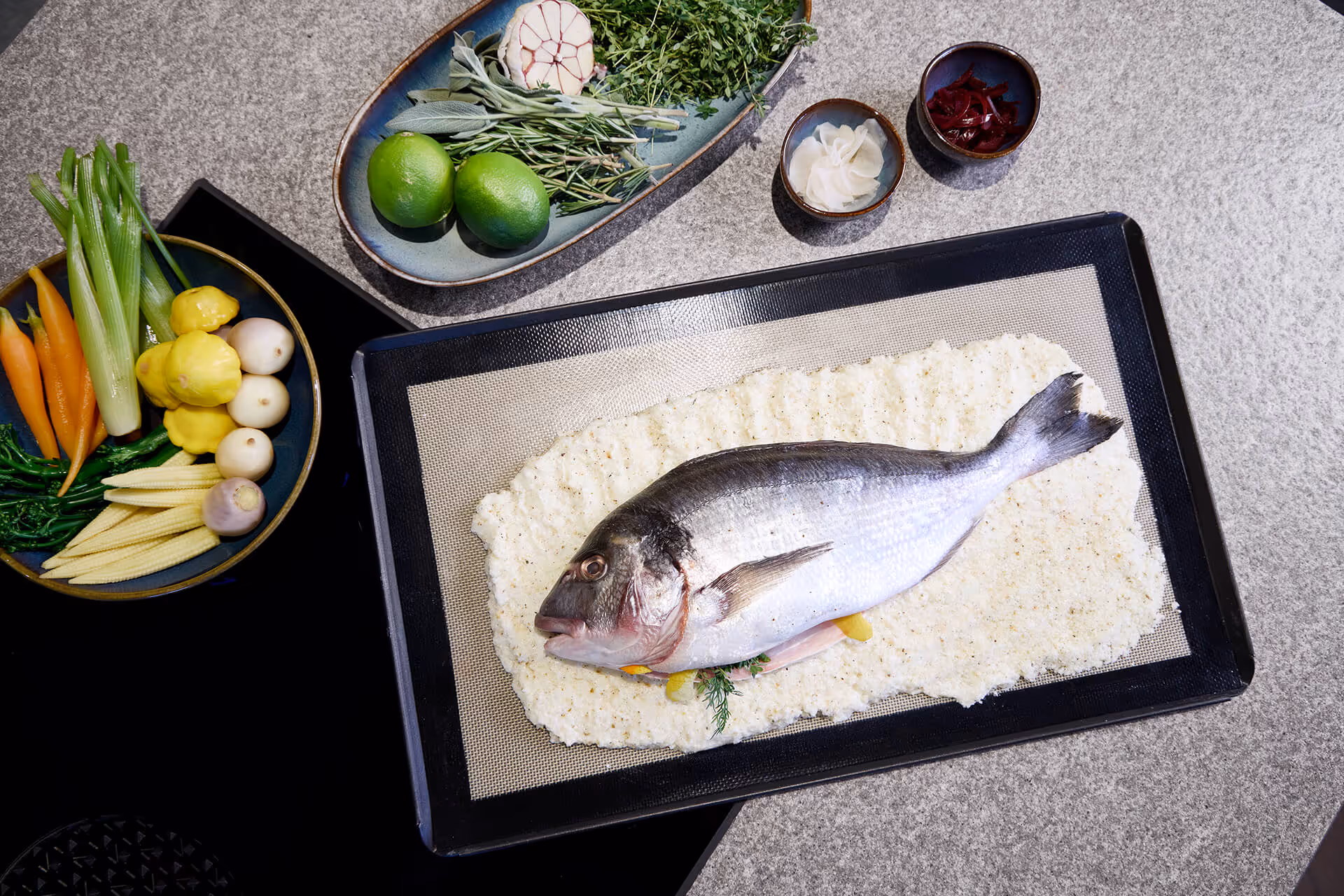 Frischer ganzer Fisch auf einem Backblech mit Salzkruste, umgeben von frischen Kräutern, Limetten und verschiedenen Gemüsesorten.