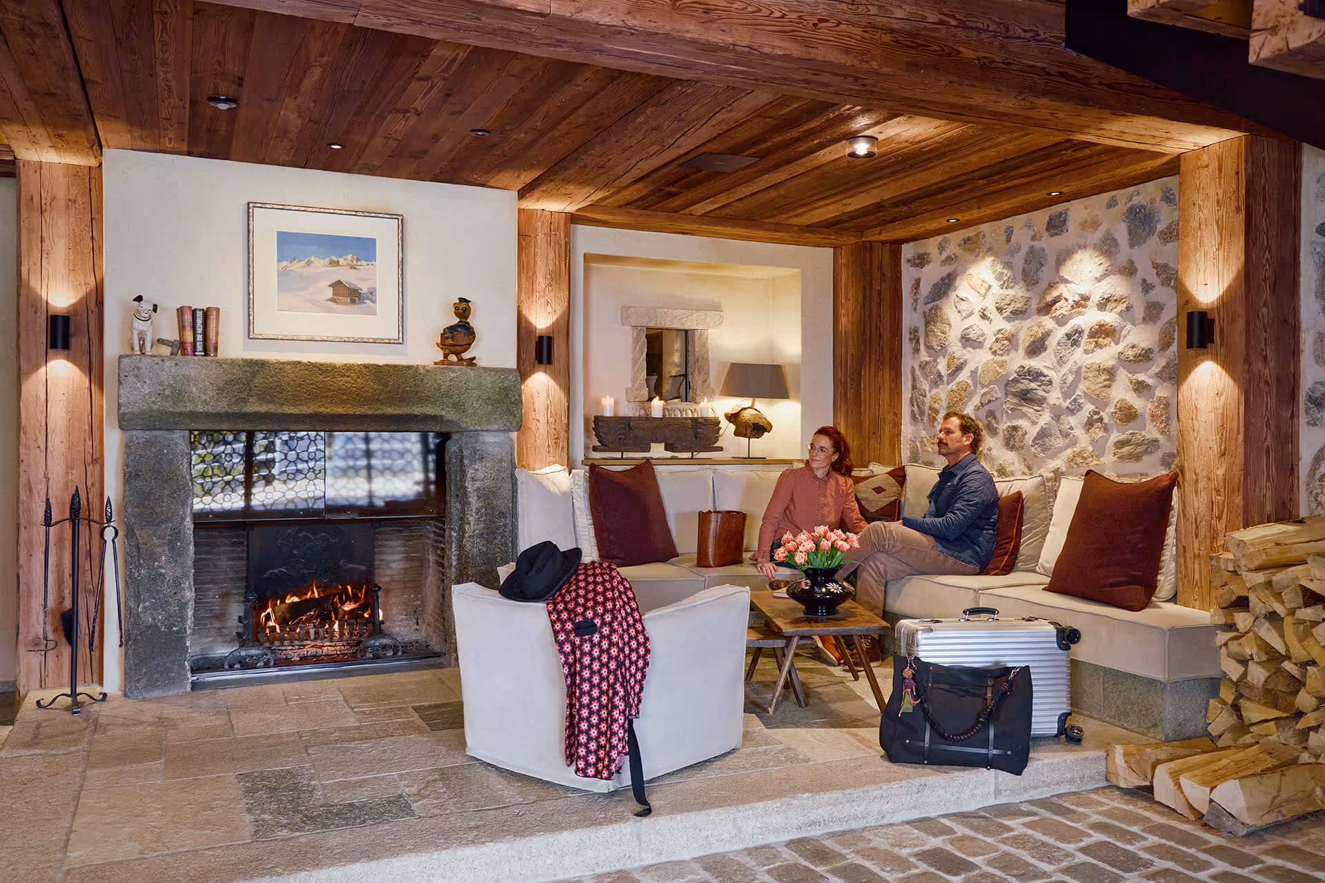 Gemütliche Wohnzimmerecke mit Steinofen, holzverkleideter Decke, zwei Personen auf einem Sofa und Reisetaschen.