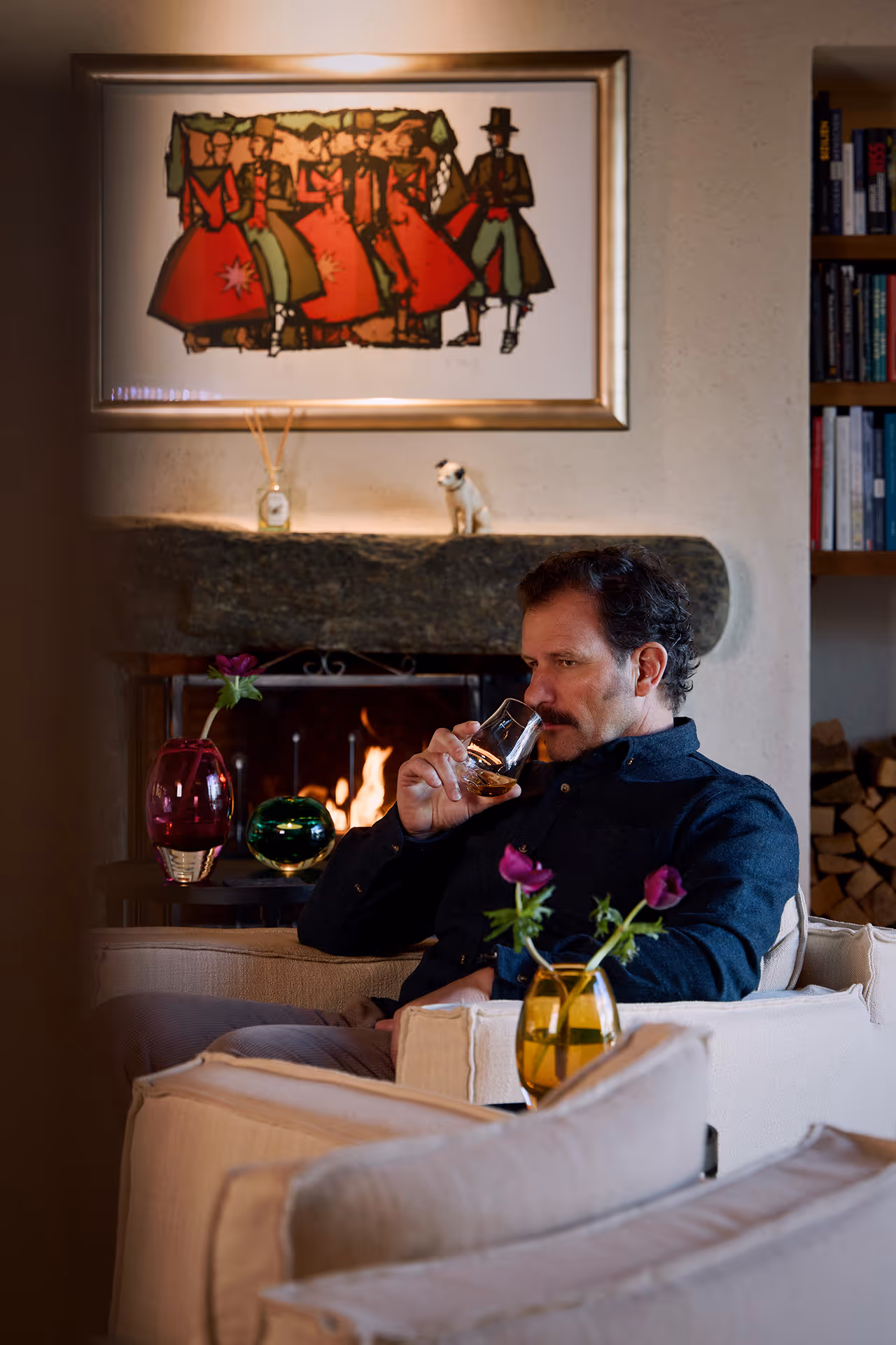Mann sitzt auf einem Sessel vor einem Kaminfeuer und riecht an einem Glas Whisky