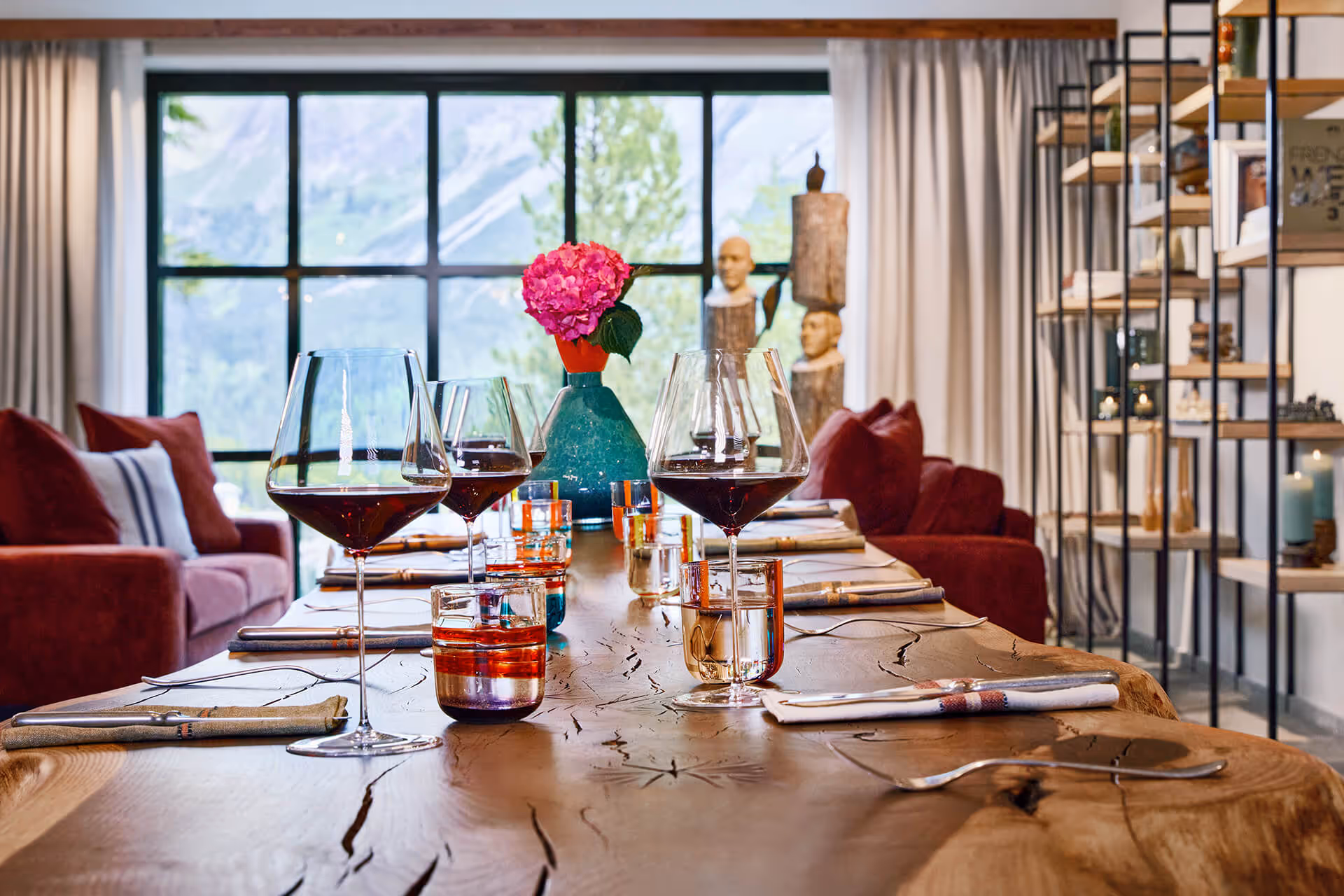 Rustikaler Holztisch mit Weingläsern und einer blauen Blumenvasen mit pinken Blüten vor grossen Fenstern mit Bergsicht
