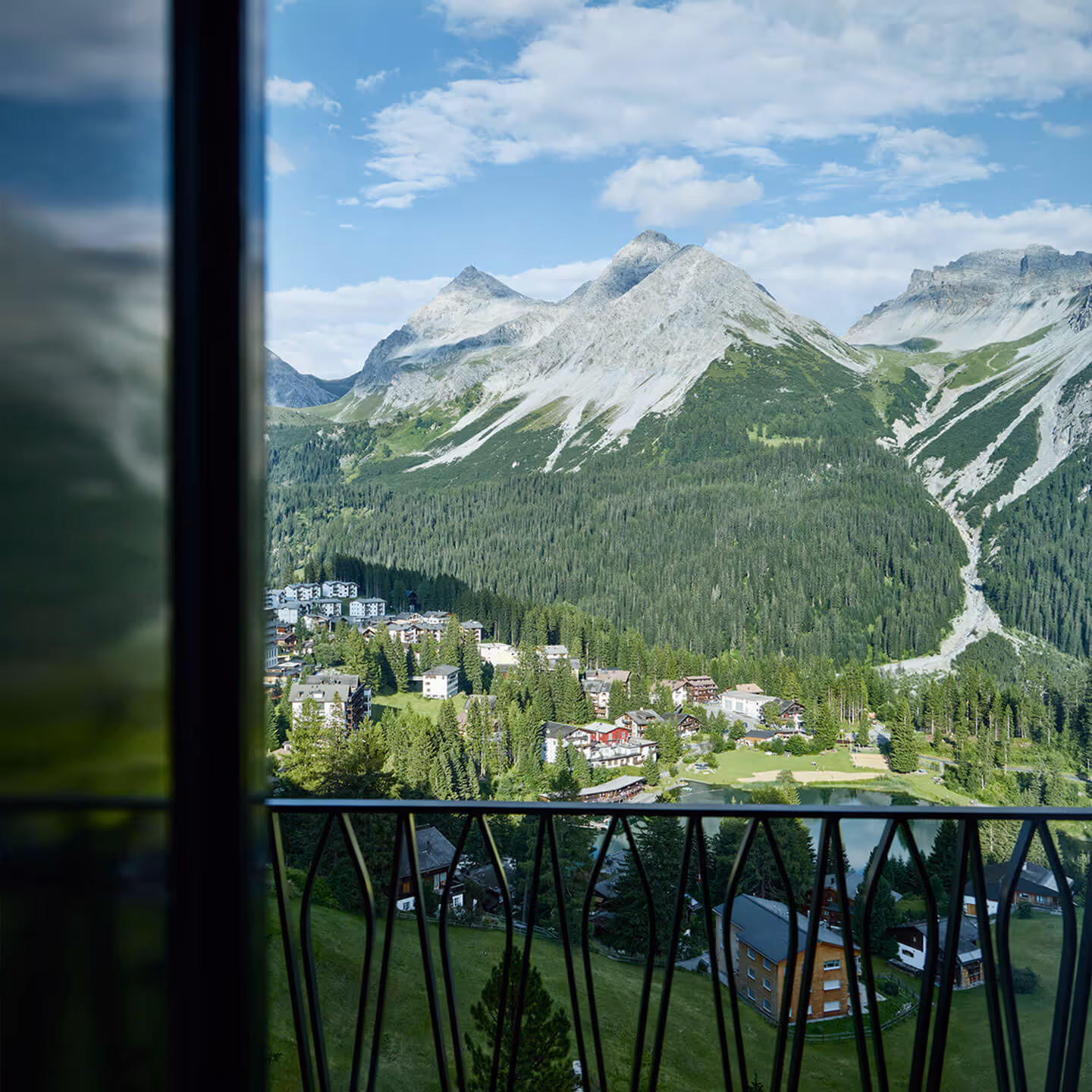 Blick von einem Balkon auf ein Alpen-Dorf mit Tannenwäldern und grasbewachsenen Hügeln vor hohen Bergen unter blauem Himmel.