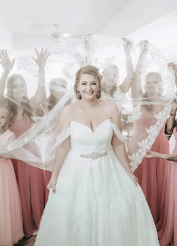 Bride smiling joyfully while bridesmaids in pink dresses hold her lace veil during wedding day preparations indoors