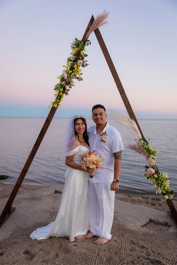 Bride and groom smiling under floral triangle arch during beachside sunset wedding ceremony