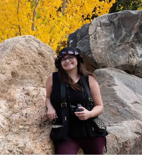 Aubrey, Shutterpix photographer and graphic designer from Denver, CO, smiling with camera in hand against mountain rocks and fall foliage