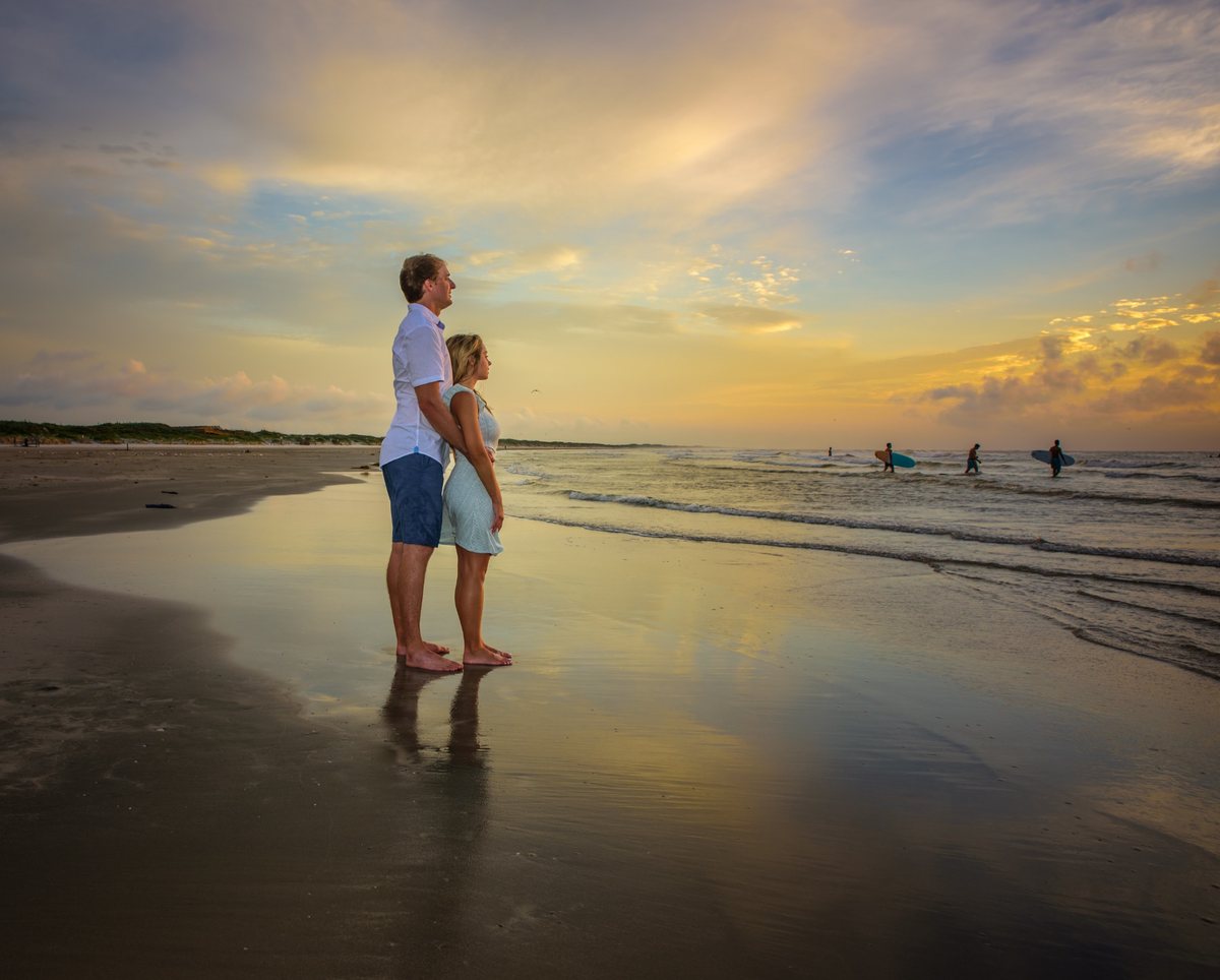 Couple standing barefoot on the wet sand of a beach at sunset, looking towards the horizon, with surfers carrying boards and waves in the background under a partly cloudy sky.
