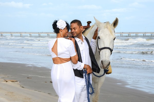 A couple dressed in black and white on a beach with a white horse, the man holding the horse's reins and the woman facing him while smiling.