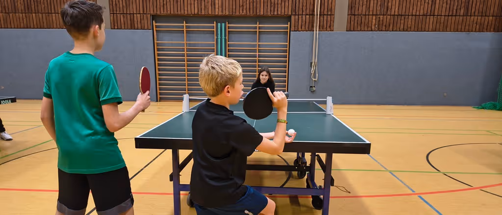 Drei Kinder spielen Tischtennis in einer Sporthalle mit Holzwänden und Turngeräten.