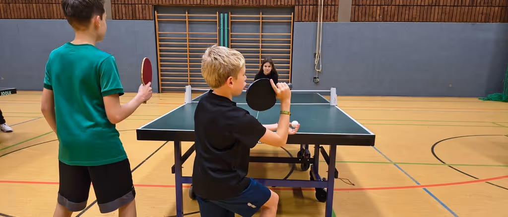 Drei Kinder spielen Tischtennis in einer Halle, zwei mit Schlägern und ein Mädchen bereit am Tisch. Das Bild zeigt unsere Tischtennis-AG.