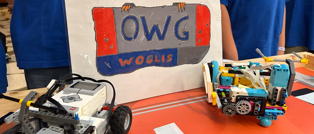 Zwei kleine programmierbare Roboter auf einem Tisch vor einem OWG Woglis Poster. Das Bild zeigt unsere Robotik AG.