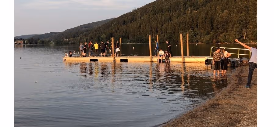 Gruppe junger Menschen am See stehen und sitzen auf einem schwimmenden Holzsteg bei Sonnenuntergang. Das Bild zeigt einen Ausschnitt der Fahrt unserer siebten Klassen nach Titisee.
