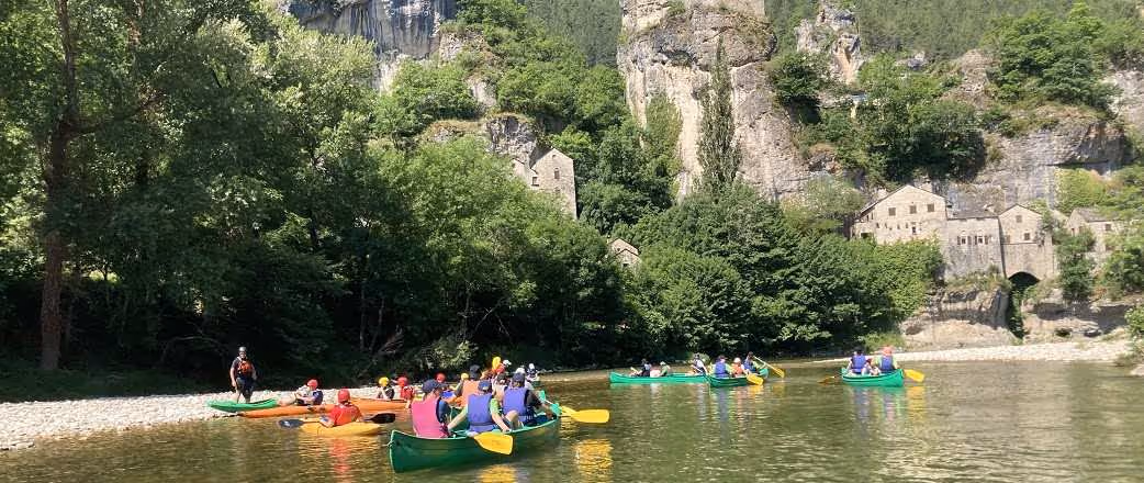 Menschen beim Kanufahren auf einem Fluss vor bewaldeten Felsen und alten Steinhäusern. Das Bild zeigt einen Ausschnitt der Exkursion an den Fluss Tarn in Südfrankreich.