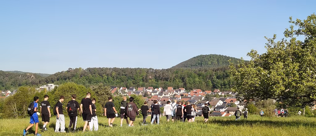 Gruppe von Menschen wandert über eine grüne Wiese mit Dorf und bewaldetem Hügel im Hintergrund. Das Bild zeigt einen Ausschnitt unseres Sponsored Walks.