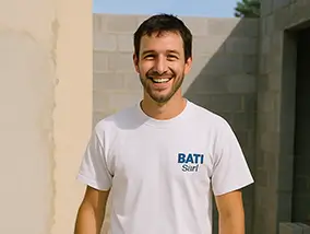 Un artisan souriant portant un t-shirt blanc devant un mur en parpaings à l'extérieur.